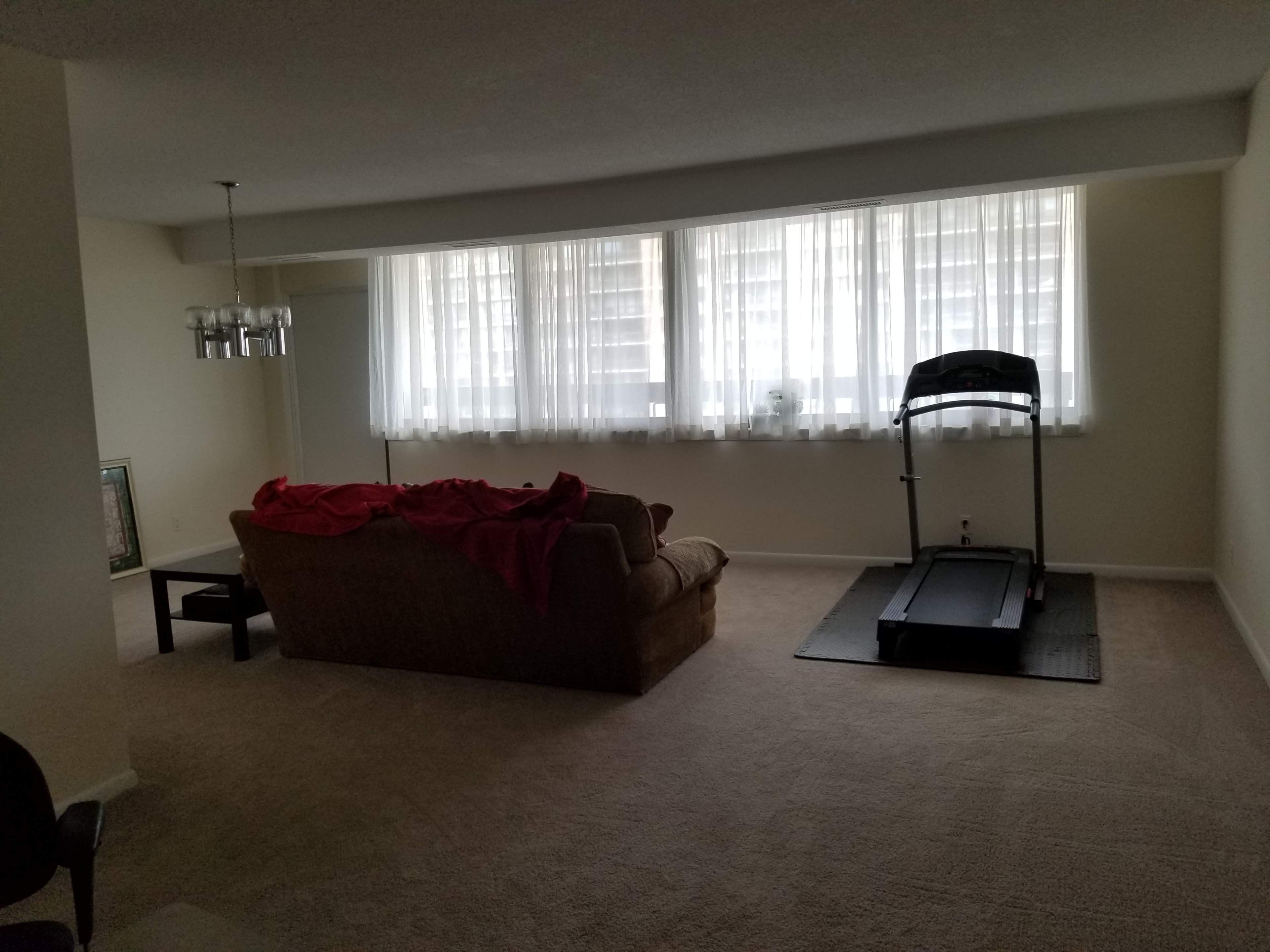 A spacious living room features a couch draped with a red blanket, a small table, and a treadmill near large windows dressed with white curtains.