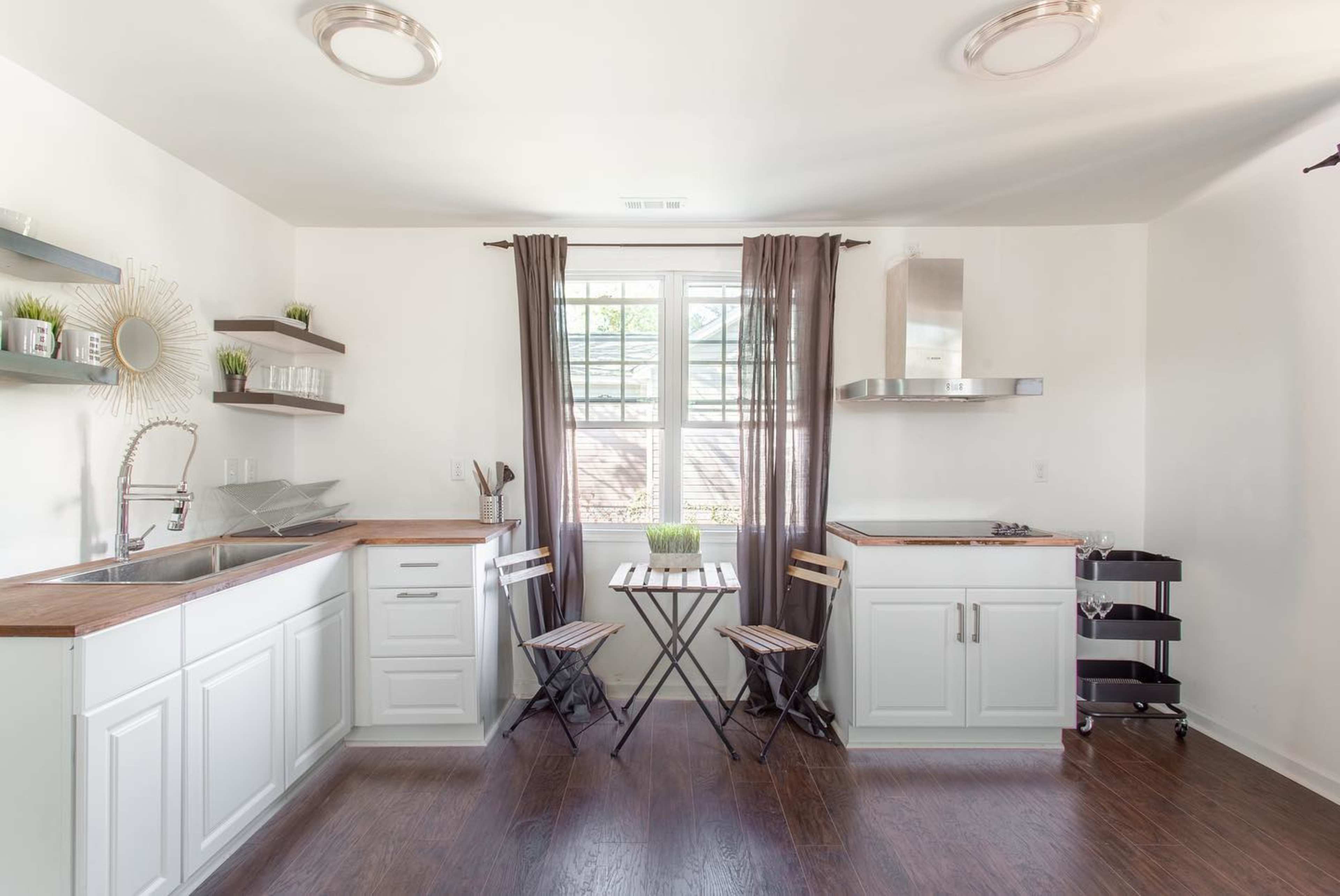 A modern kitchen with white cabinetry, wooden countertops, a window with curtains, and a small dining table with chairs in the corner.
