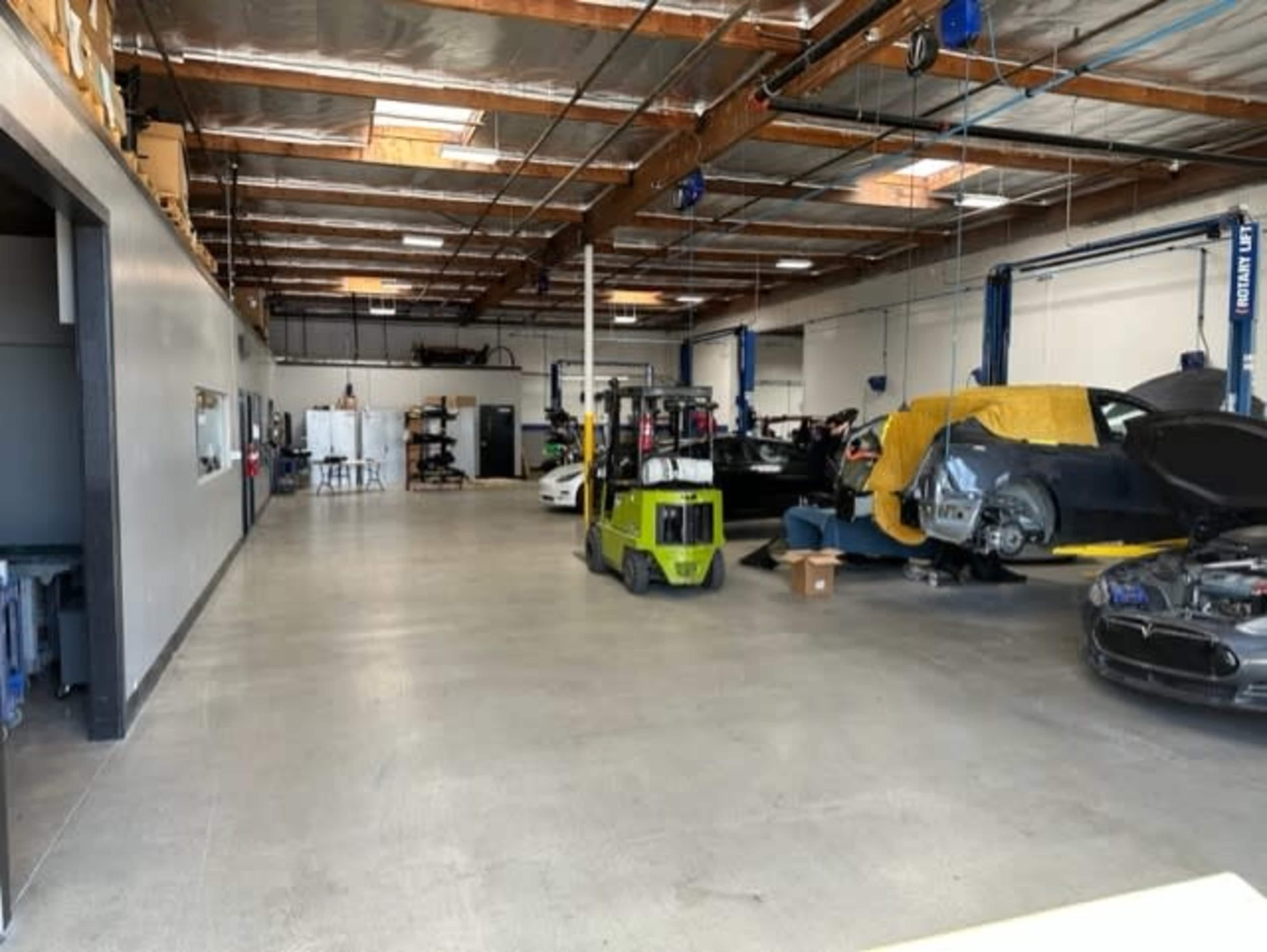 A spacious automotive workshop with various cars, a yellow forklift, and equipment scattered throughout the area.
