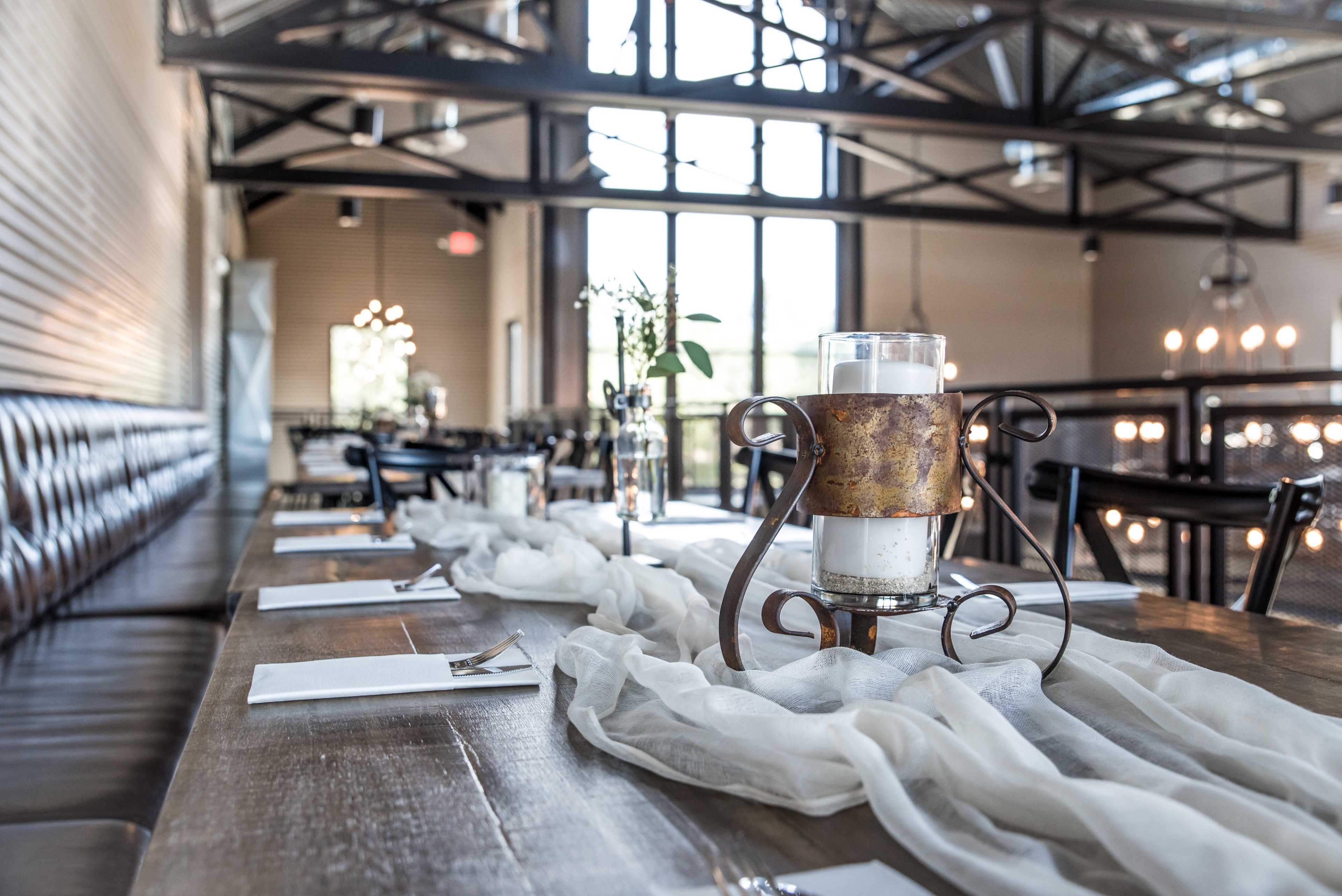 A long dining table is set with white linens and simple centerpieces in a well-lit industrial-style restaurant.