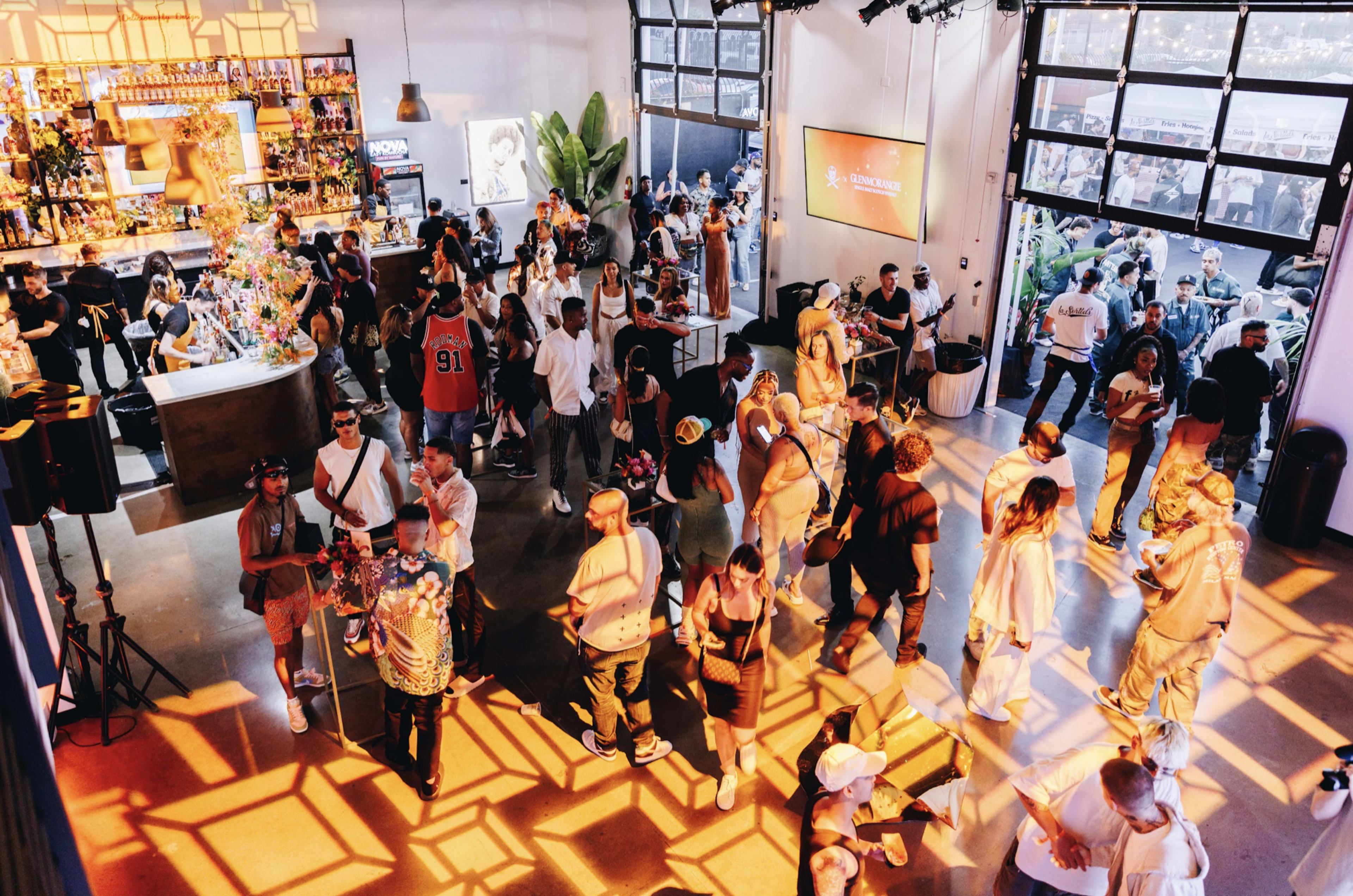 The image shows a crowded indoor event space with people mingling and enjoying a vibrant atmosphere, while sunlight casts patterned shadows on the floor.