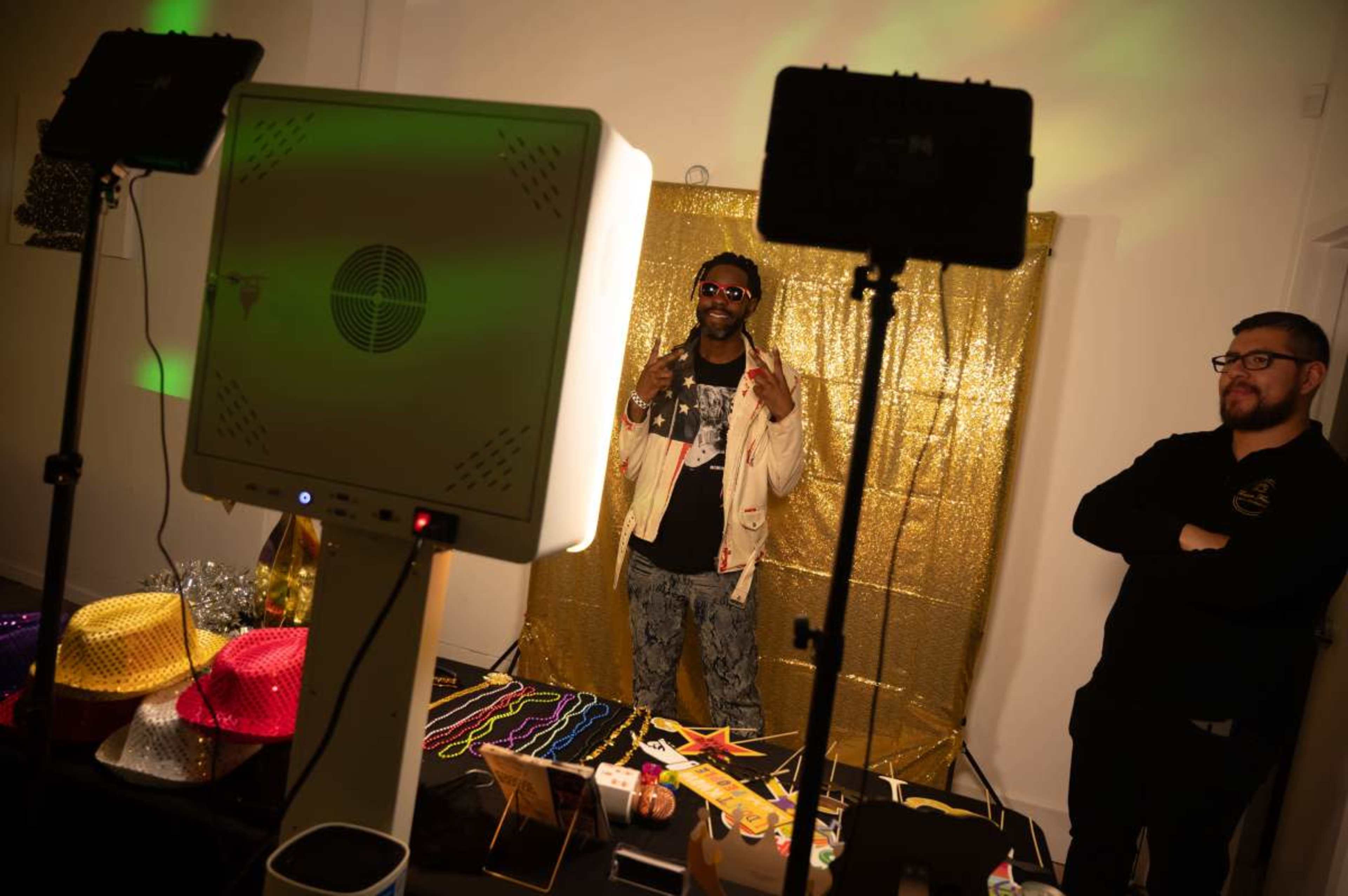 A man poses with peace signs in front of a glittery backdrop while another man stands to the side, observing.