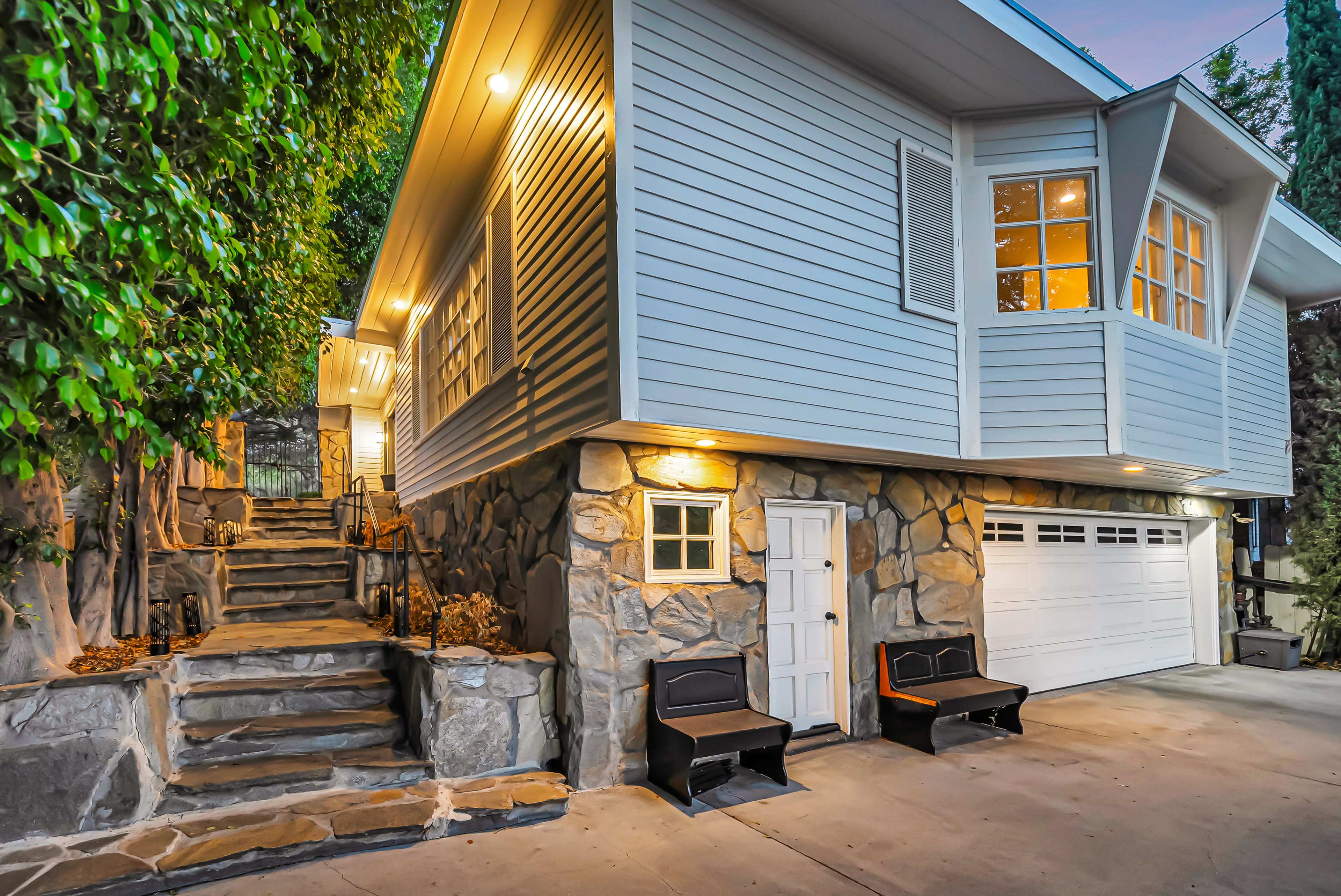 A modern house with a stone foundation has a set of stairs leading up to an entrance, and features a garage and two black benches in front.