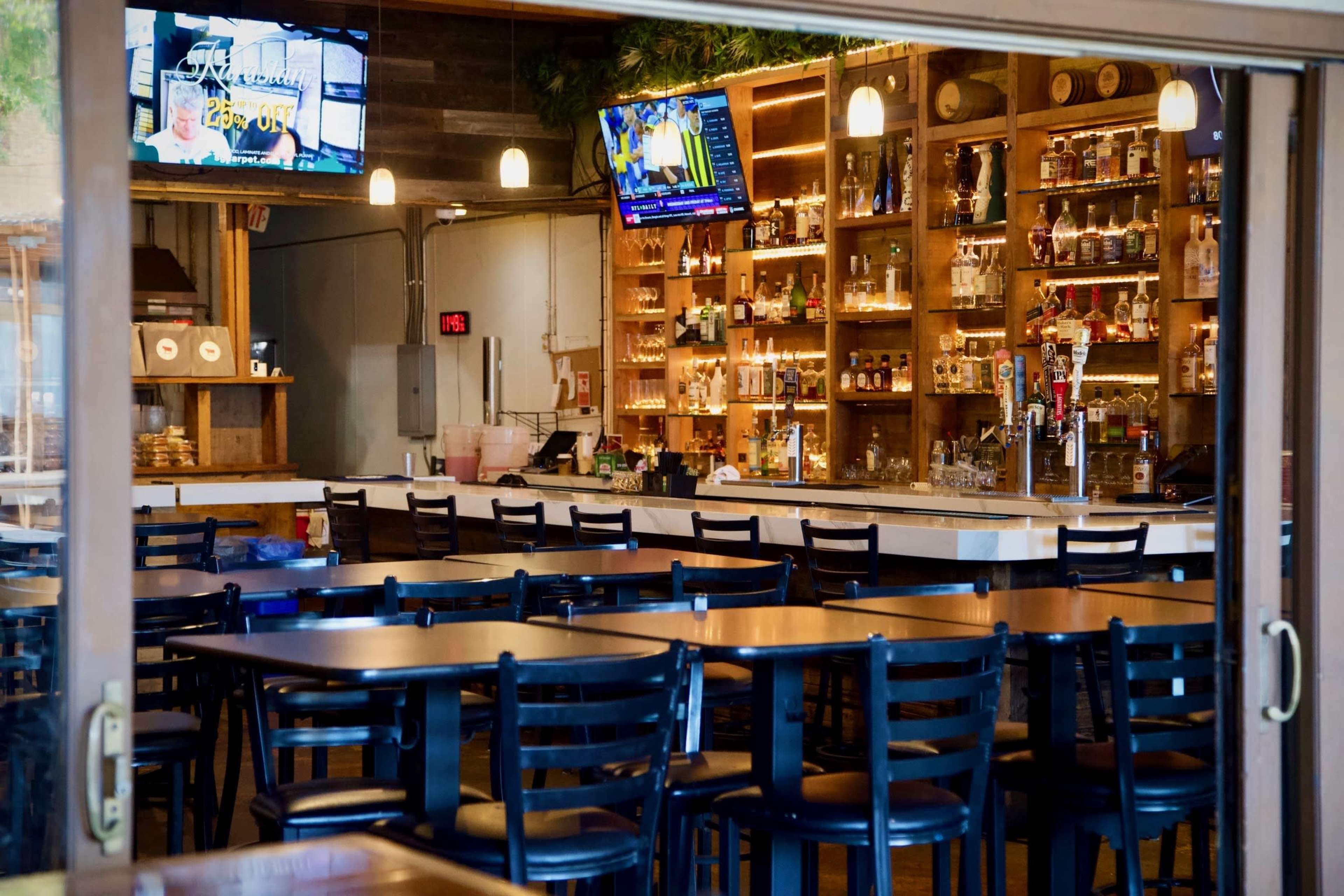 The image shows a bar and dining area with wooden tables, black chairs, and shelves lined with various bottles, illuminated by two televisions.