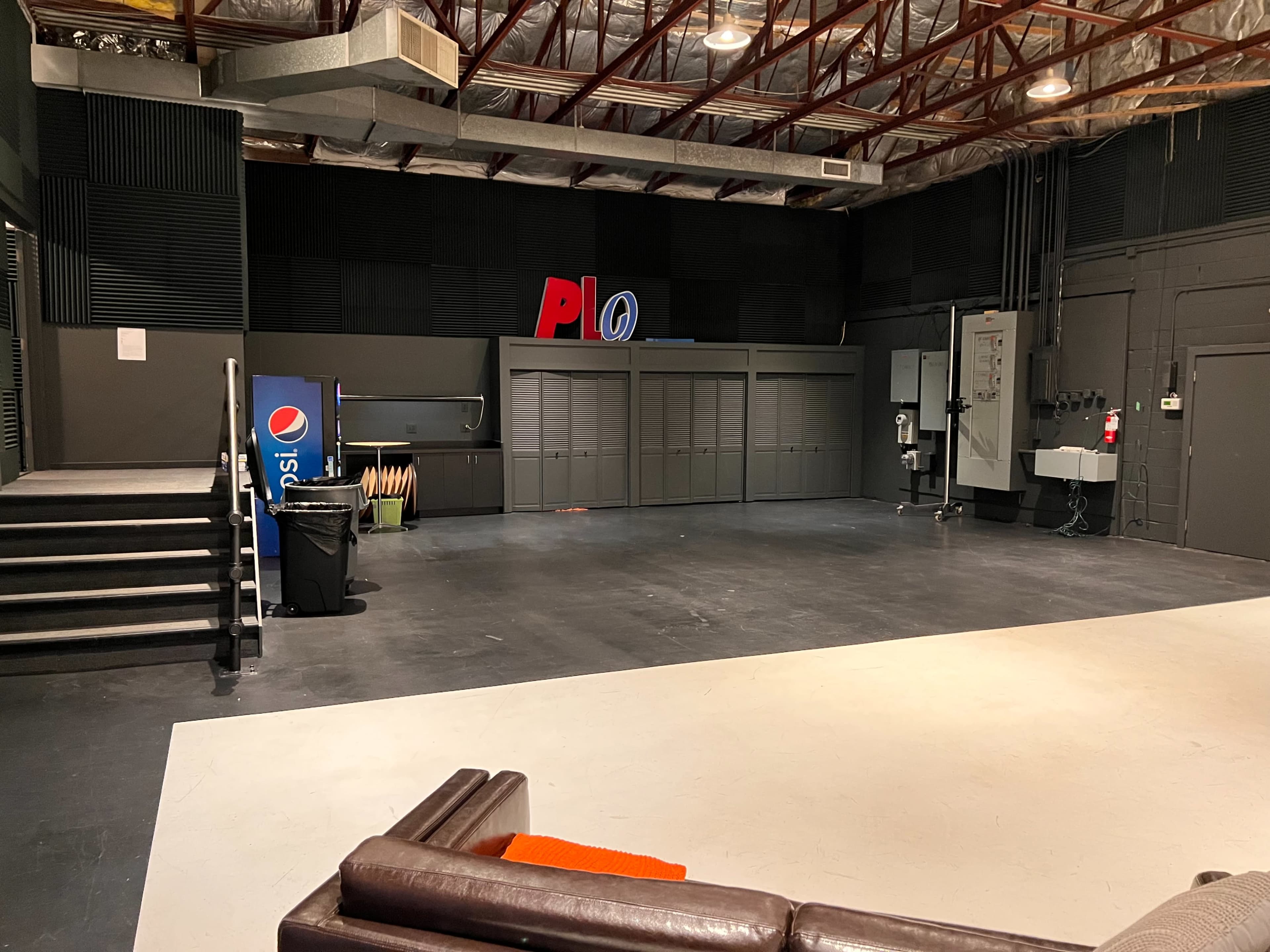An empty studio space with dark walls, a white floor, and various equipment and signage along the back wall.