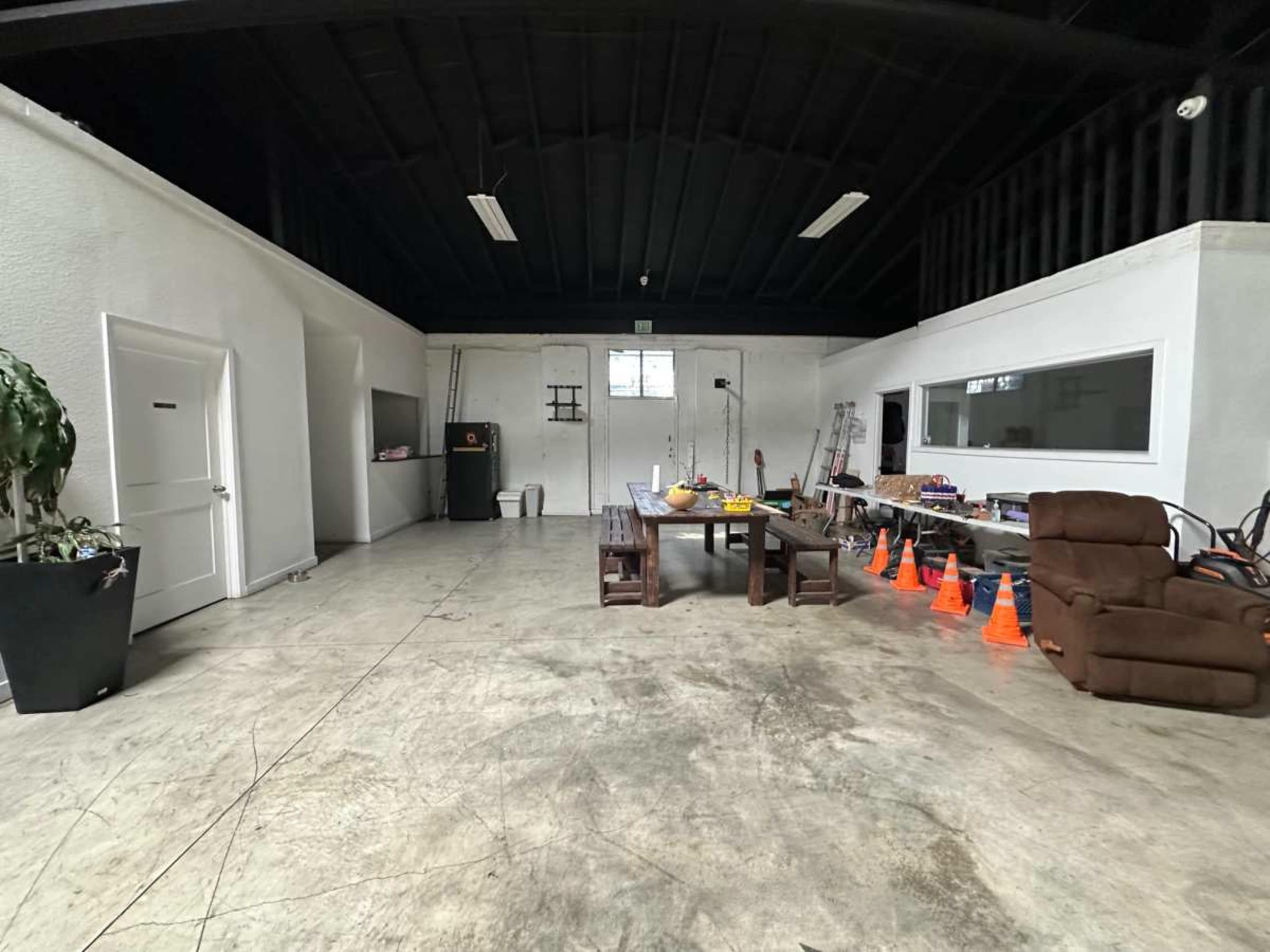The image shows a spacious, empty room with a table, chairs, and a few pieces of furniture, including a recliner and traffic cones arranged along one side.