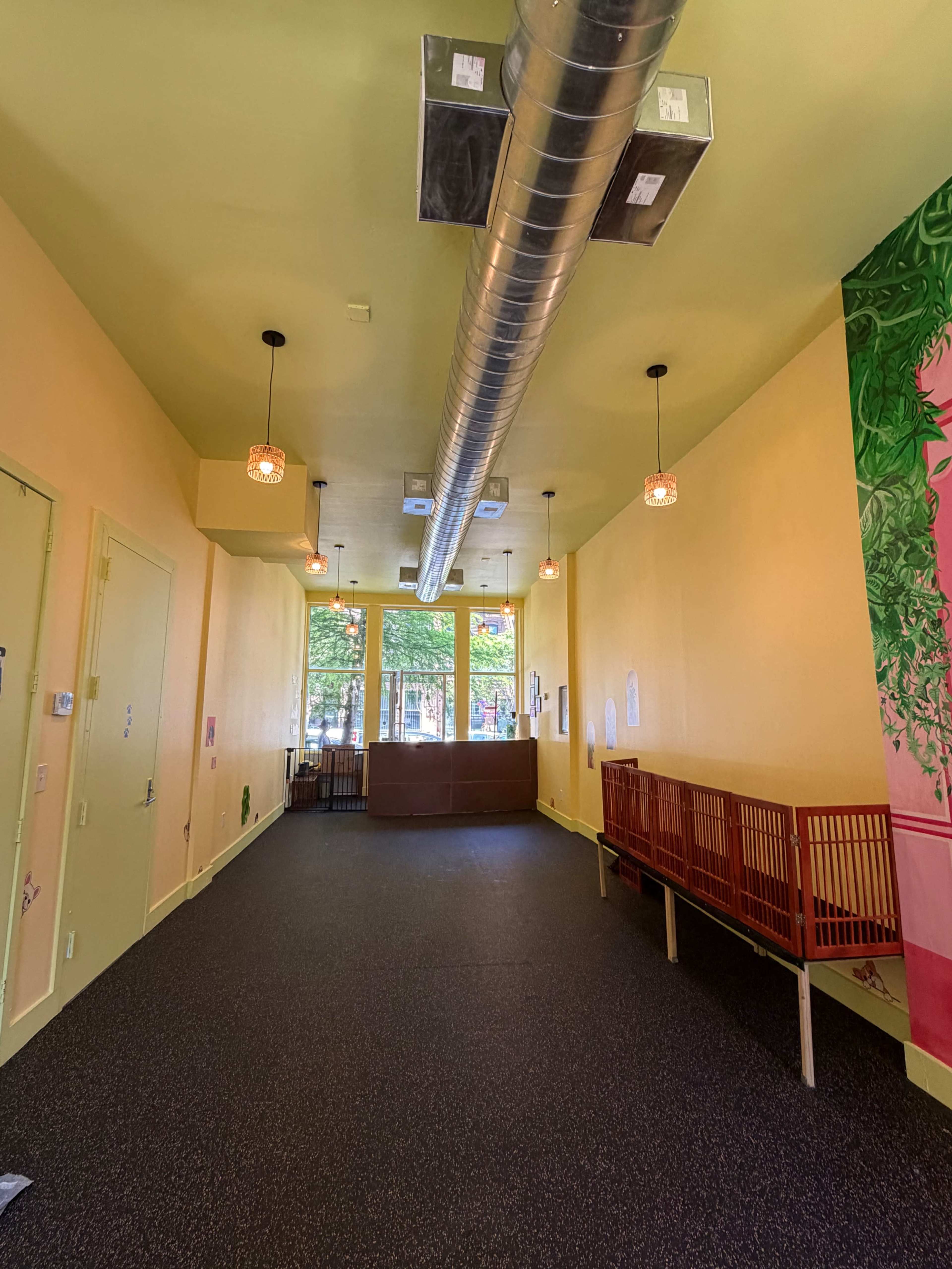 The image shows the interior of a bright, empty room with yellow walls, a ceiling vent, and wooden benches along one side.