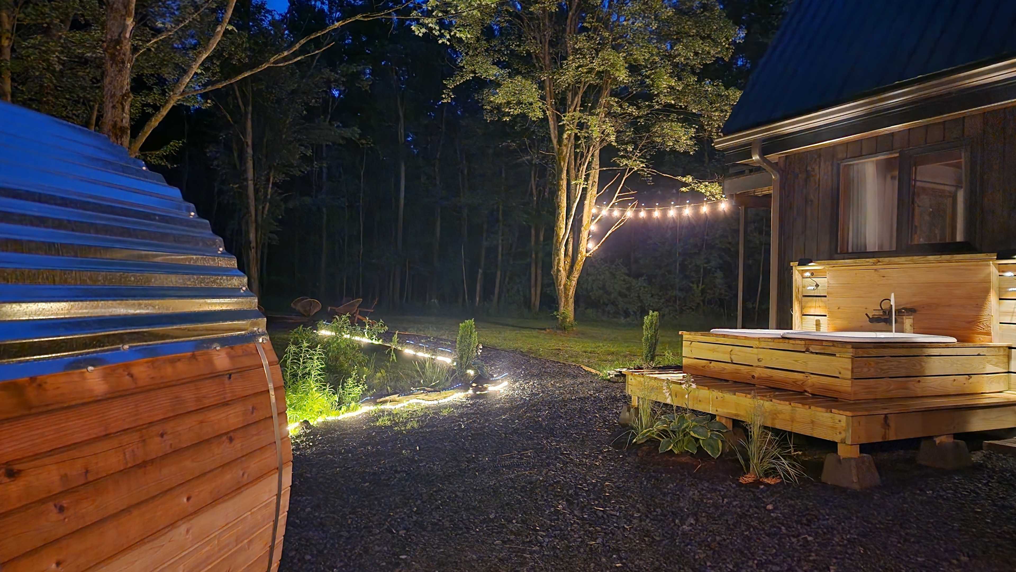 The scene shows a nighttime outdoor area featuring a wooden hot tub, a pathway illuminated by lights, and a tree adorned with string lights.
