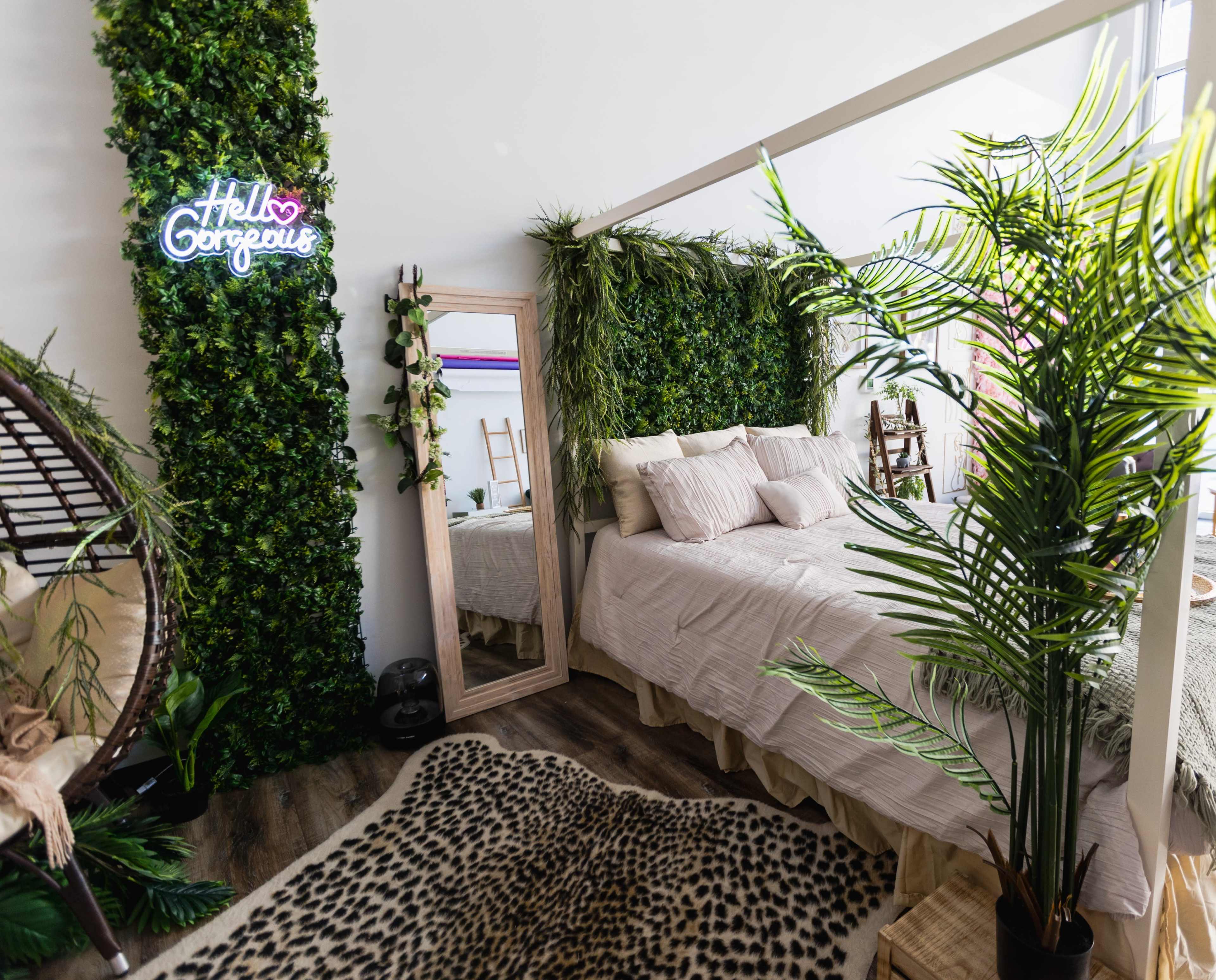 A bedroom features a bed surrounded by greenery, a large mirror, and a neon sign that says "Hello Gorgeous."