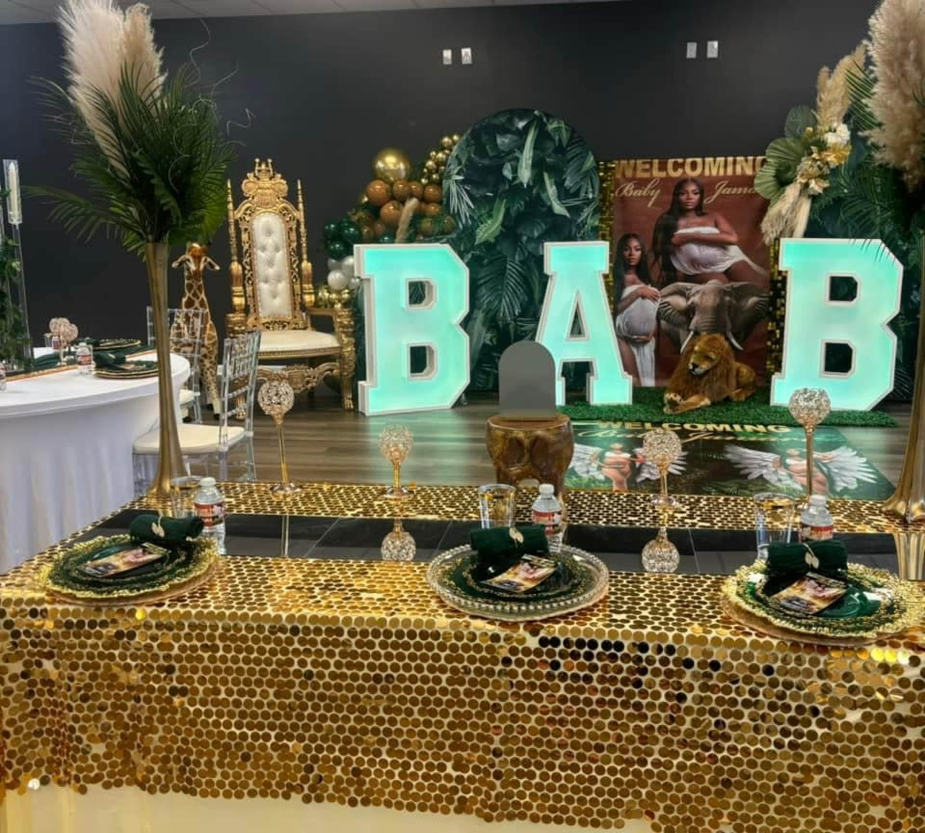 The image shows a decorated event space featuring a gold sequined tablecloth, large illuminated letters spelling "BAB," and tropical-themed decor with greenery and balloons.
