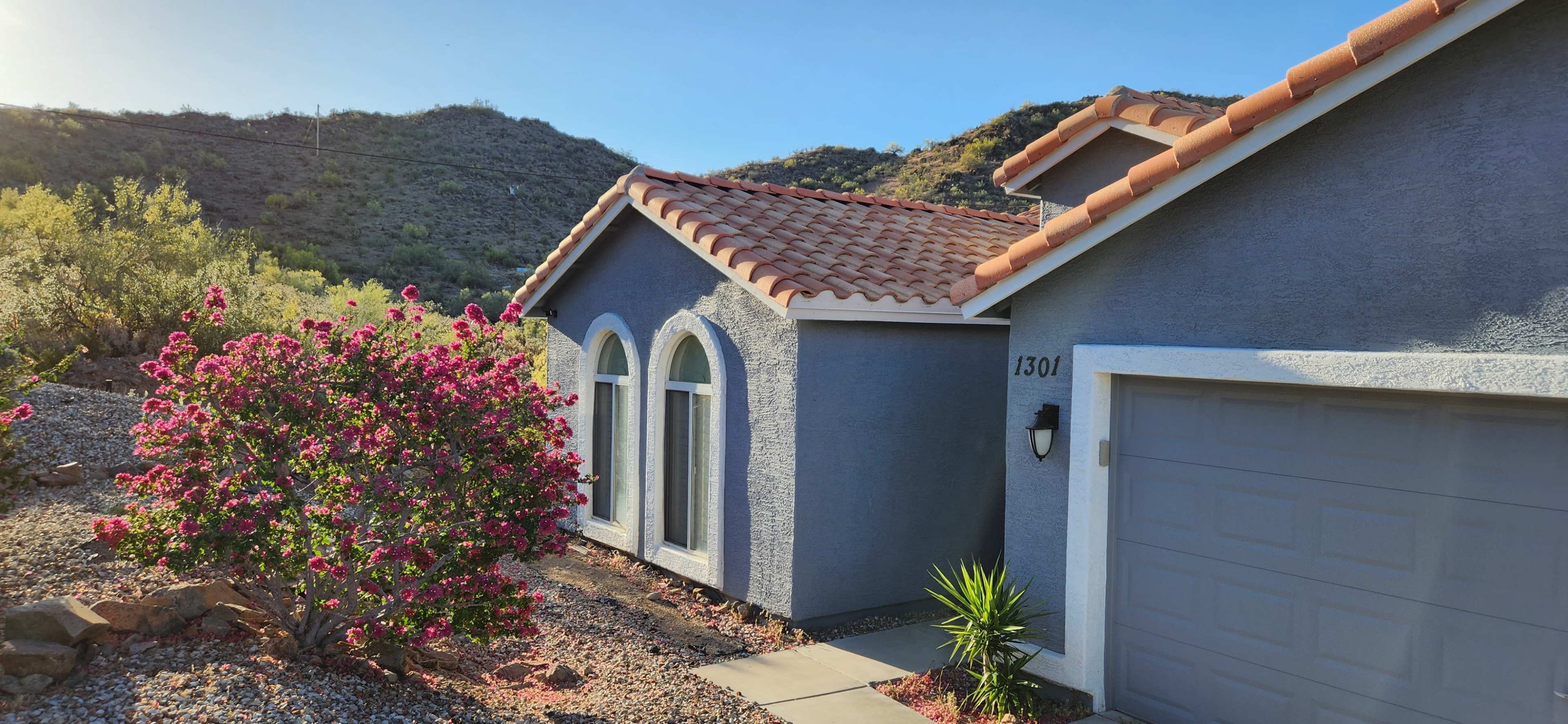 A house with a tiled roof and arched windows is situated next to a flowering bush and a garage, against a backdrop of rolling hills.