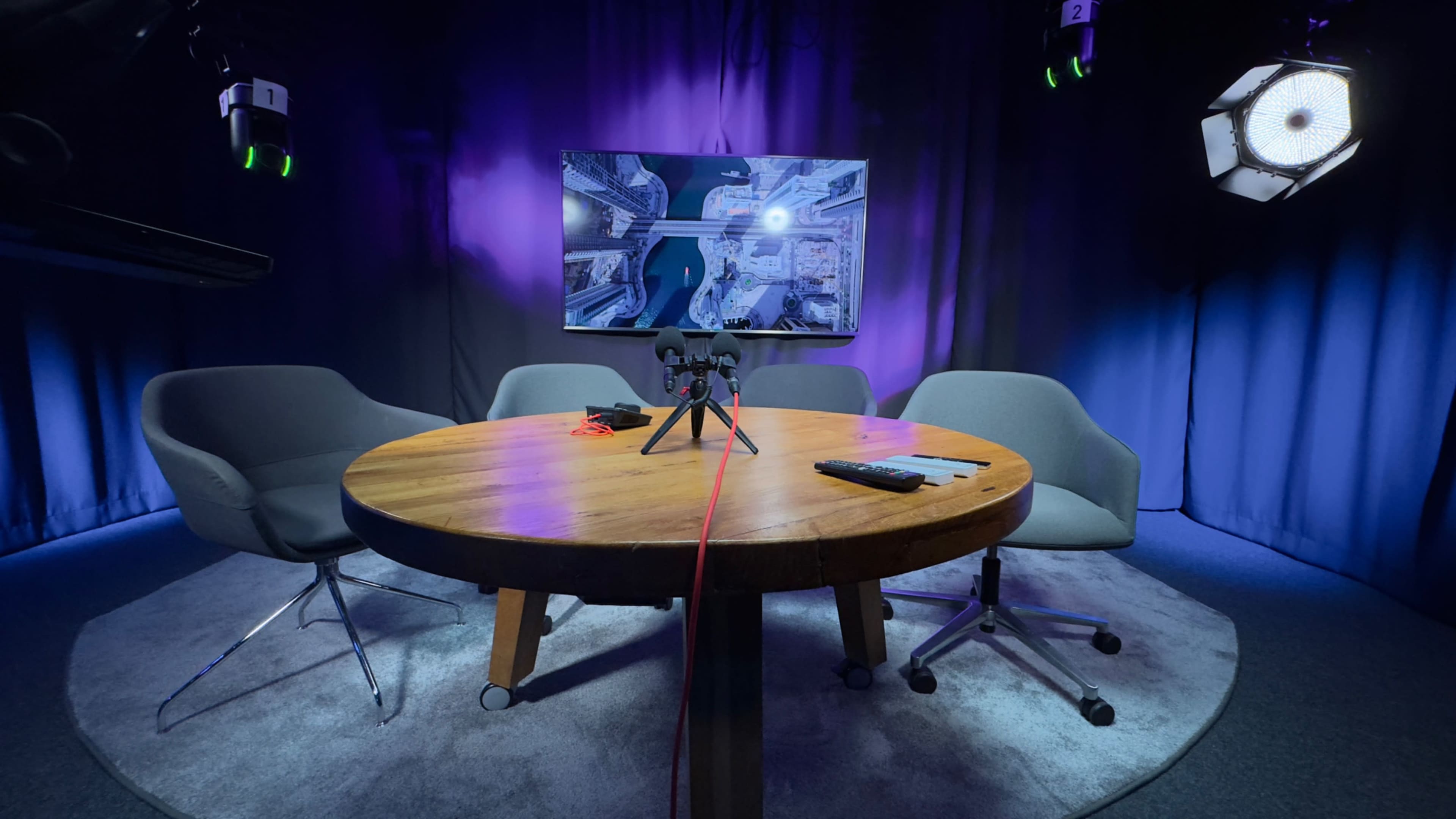 A modern studio setup features a round wooden table with three gray chairs, a camera on a tripod, and a large screen displaying an aerial view of a city.