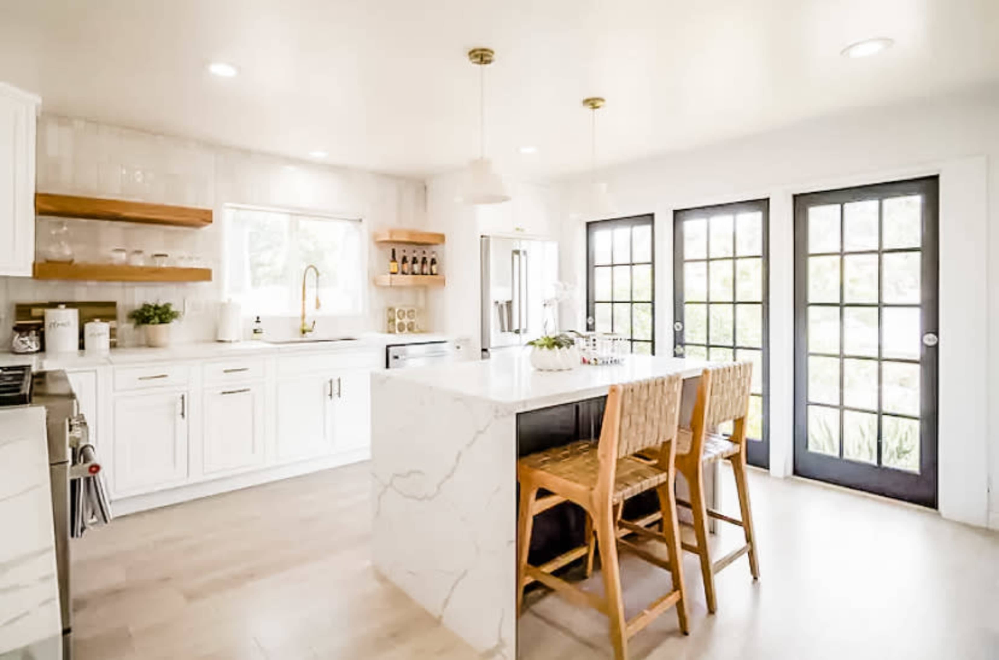 A modern kitchen features white cabinetry, a large marble island with three wooden stools, and glass-paned doors leading outside.