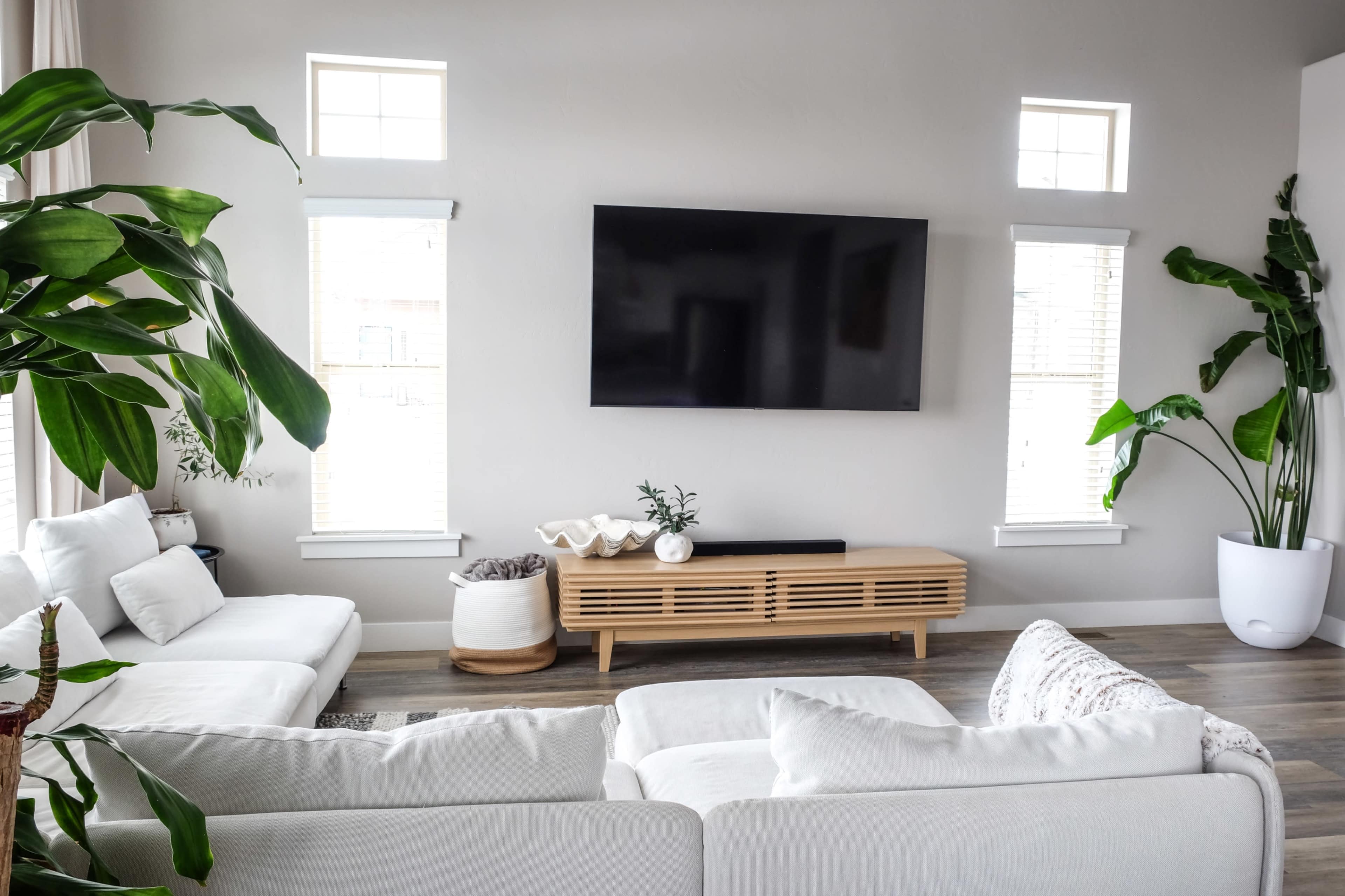 The image shows a modern living room with a large flat-screen TV mounted on a light gray wall, flanked by two windows, and features a sectional sofa and a wooden media console.