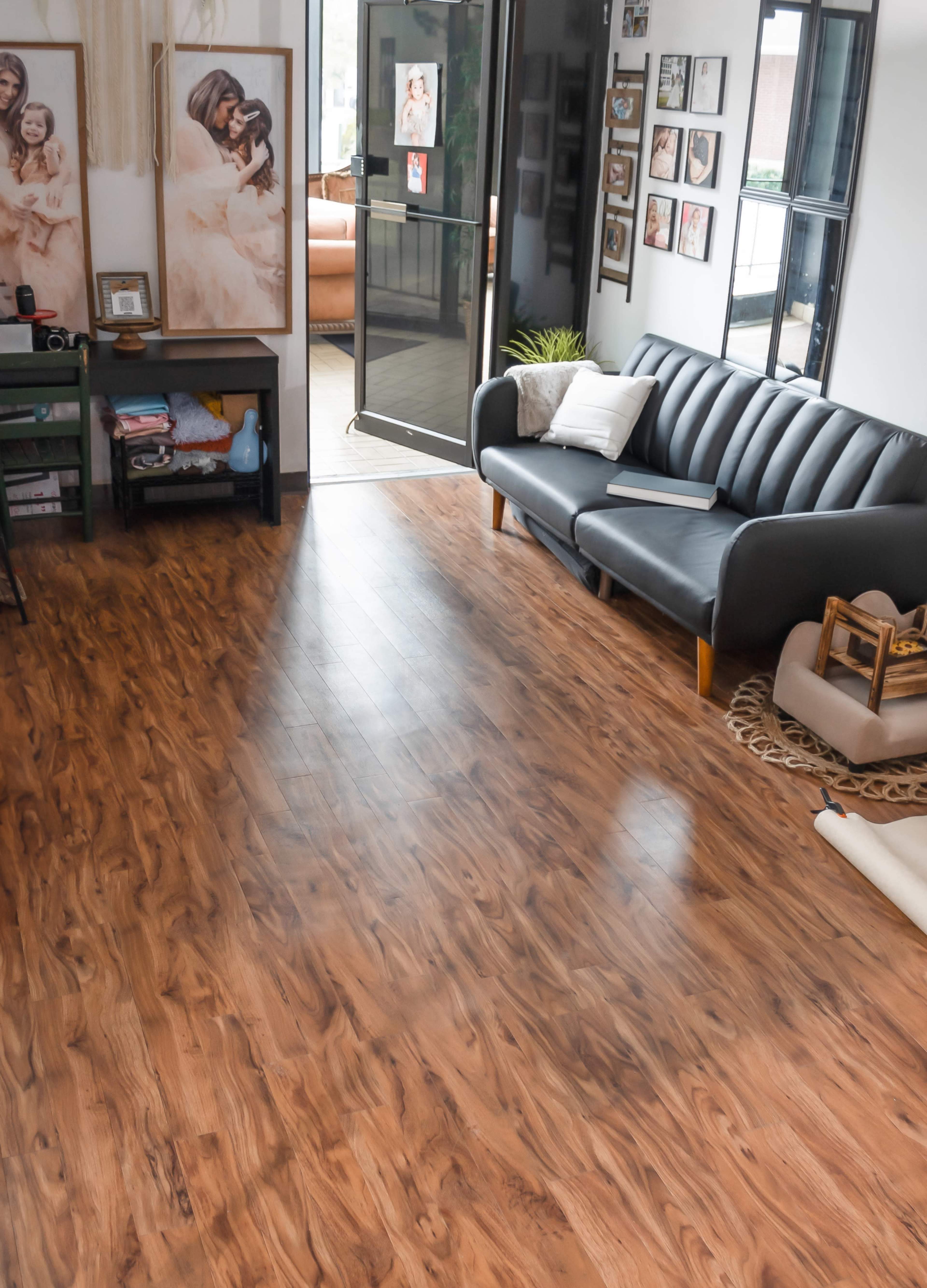 A modern living room with a black leather sofa, wooden flooring, and large windows letting in natural light.