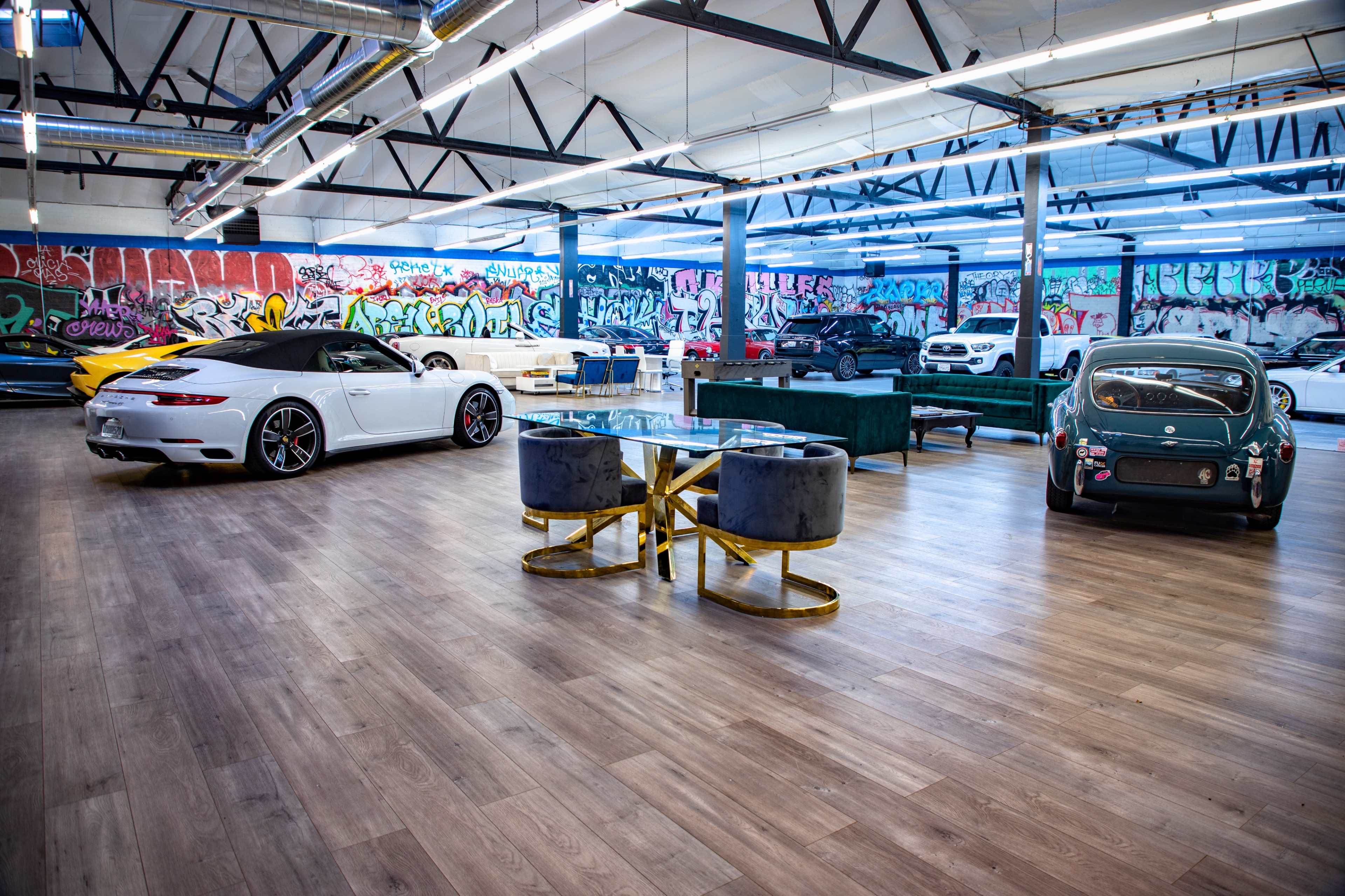 The image shows a modern showroom featuring a white sports car, a vintage green car, and elegant furniture against a backdrop of colorful graffiti on the walls.