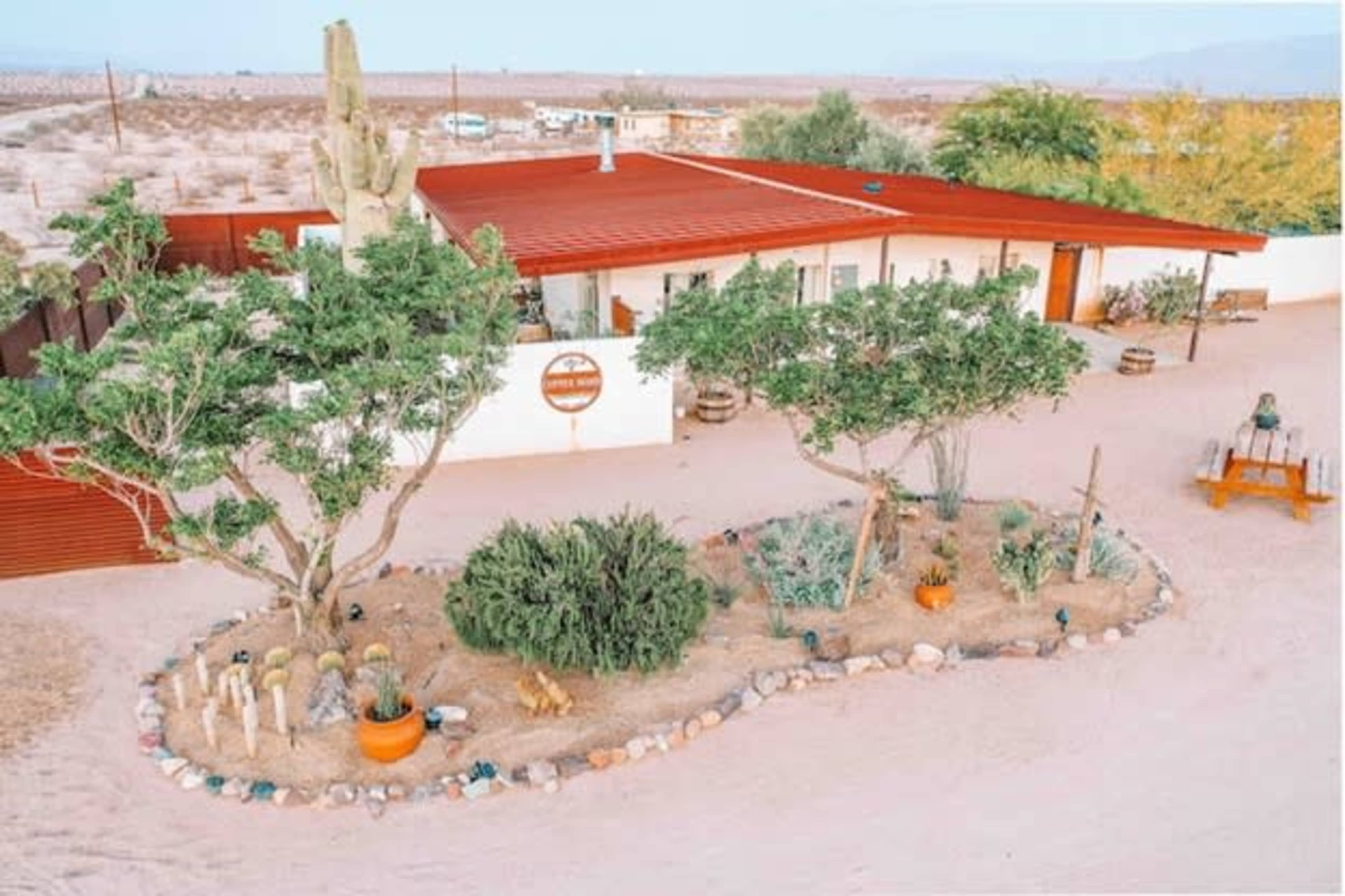 A single-story house with a red metal roof is surrounded by desert landscaping featuring cacti, shrubs, and decorative stones.