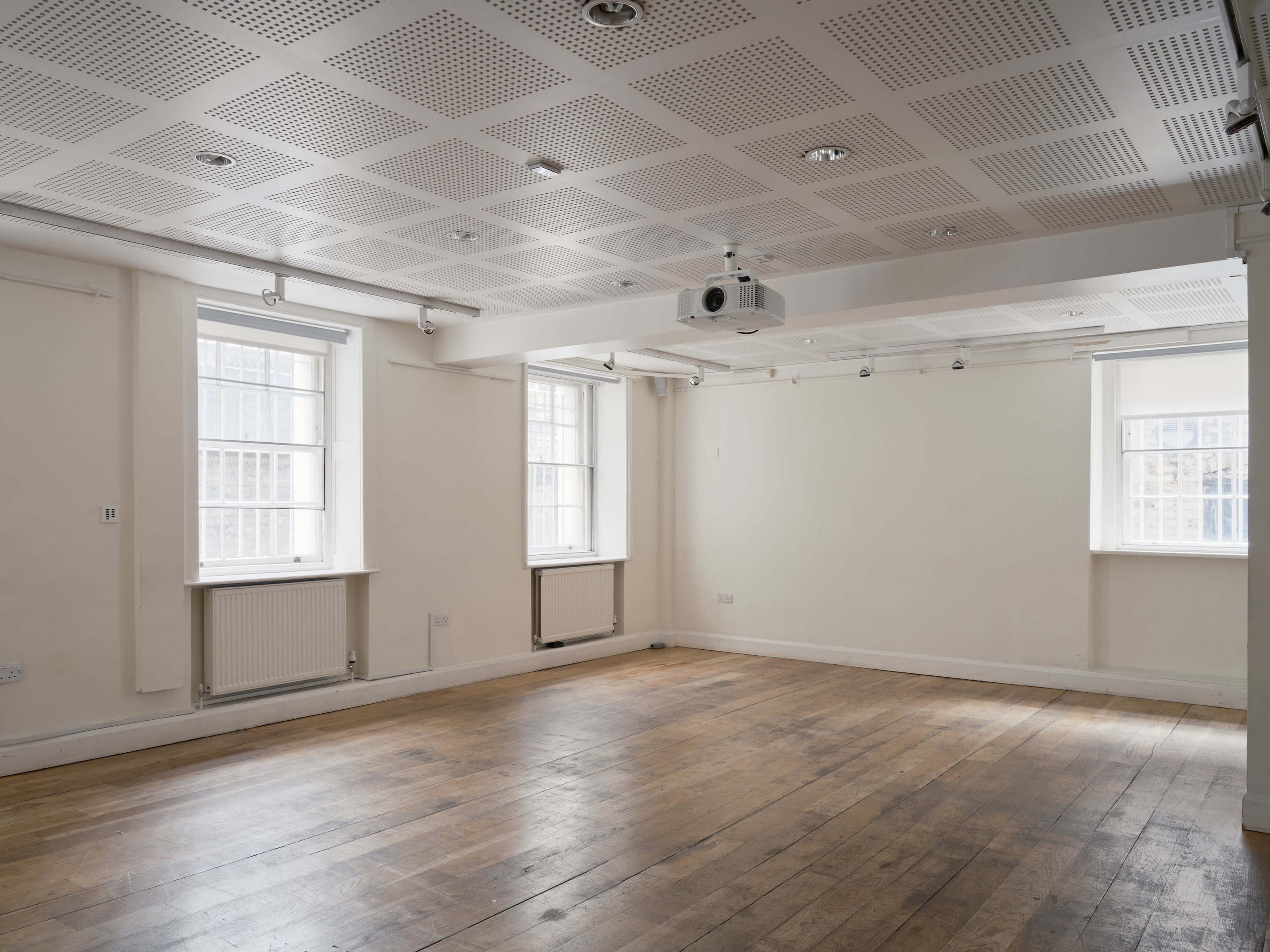 The image shows an empty room with wooden flooring, two windows, and a ceiling-mounted projector.