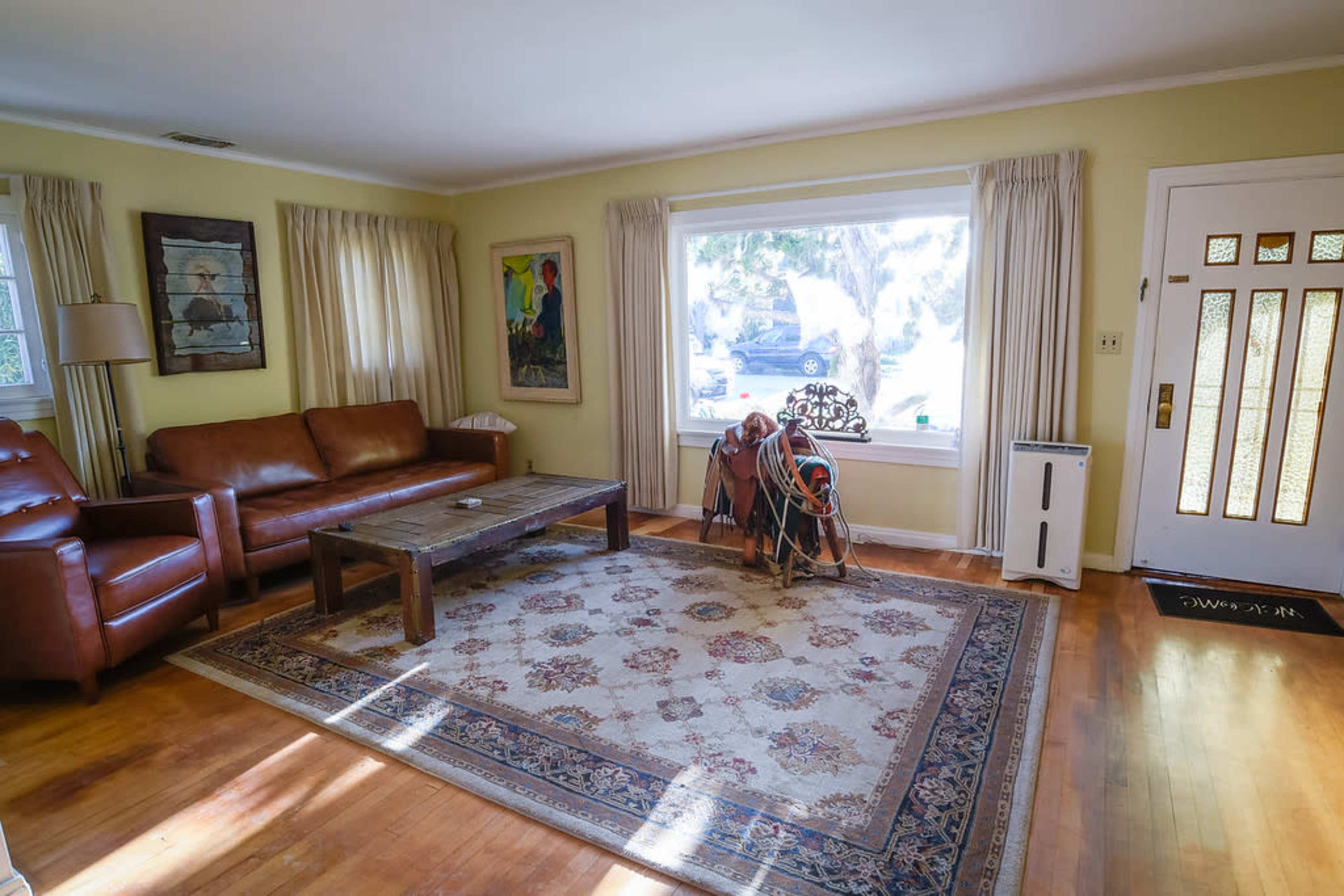 A living room features a brown leather sofa, a wooden coffee table, and a decorative rug, with a large window and an entrance door visible.