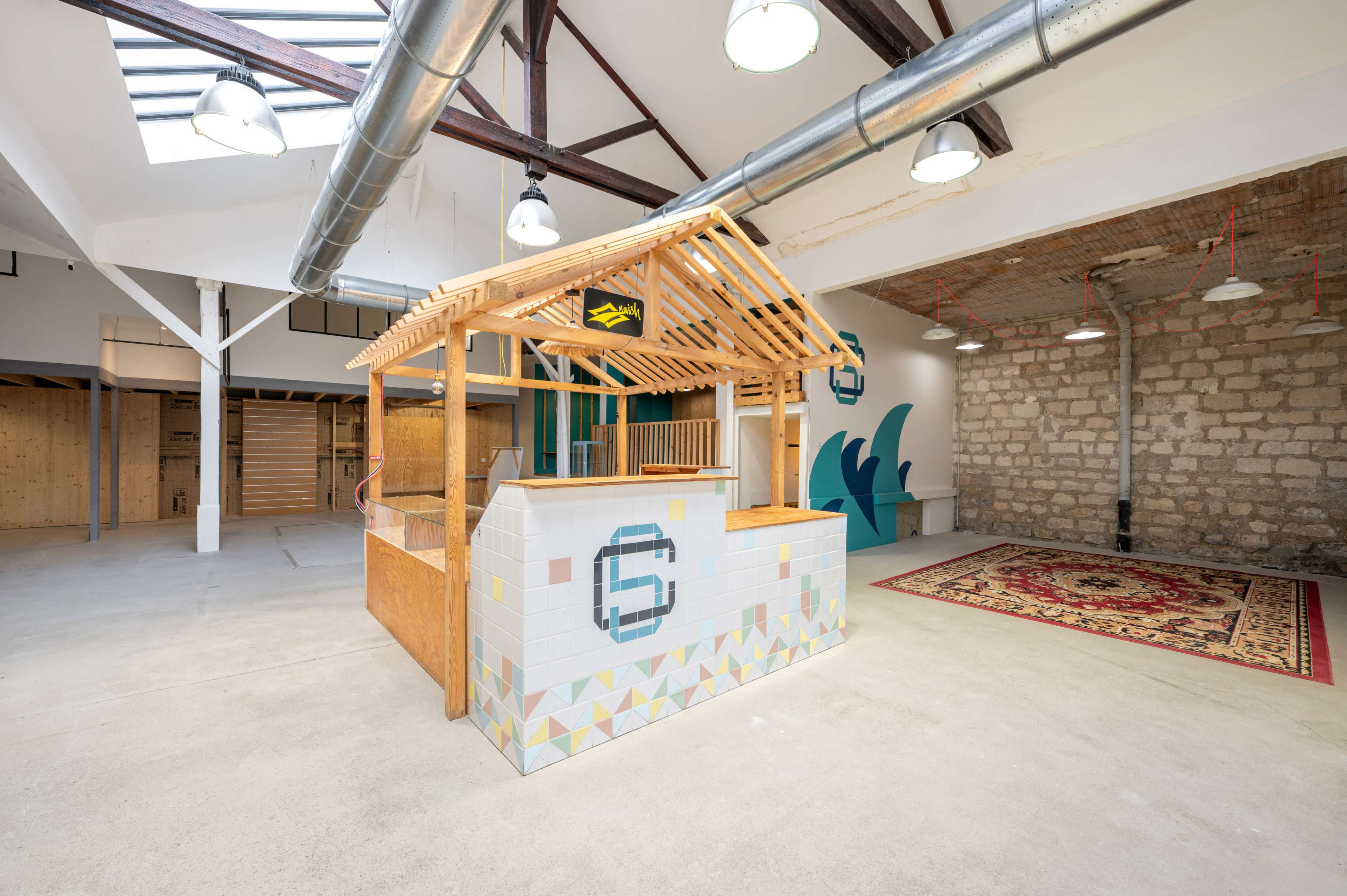 A spacious, minimalist interior featuring a wooden structure resembling a reception or kiosk area, with a patterned tiled base and exposed industrial elements like pipes and beams.
