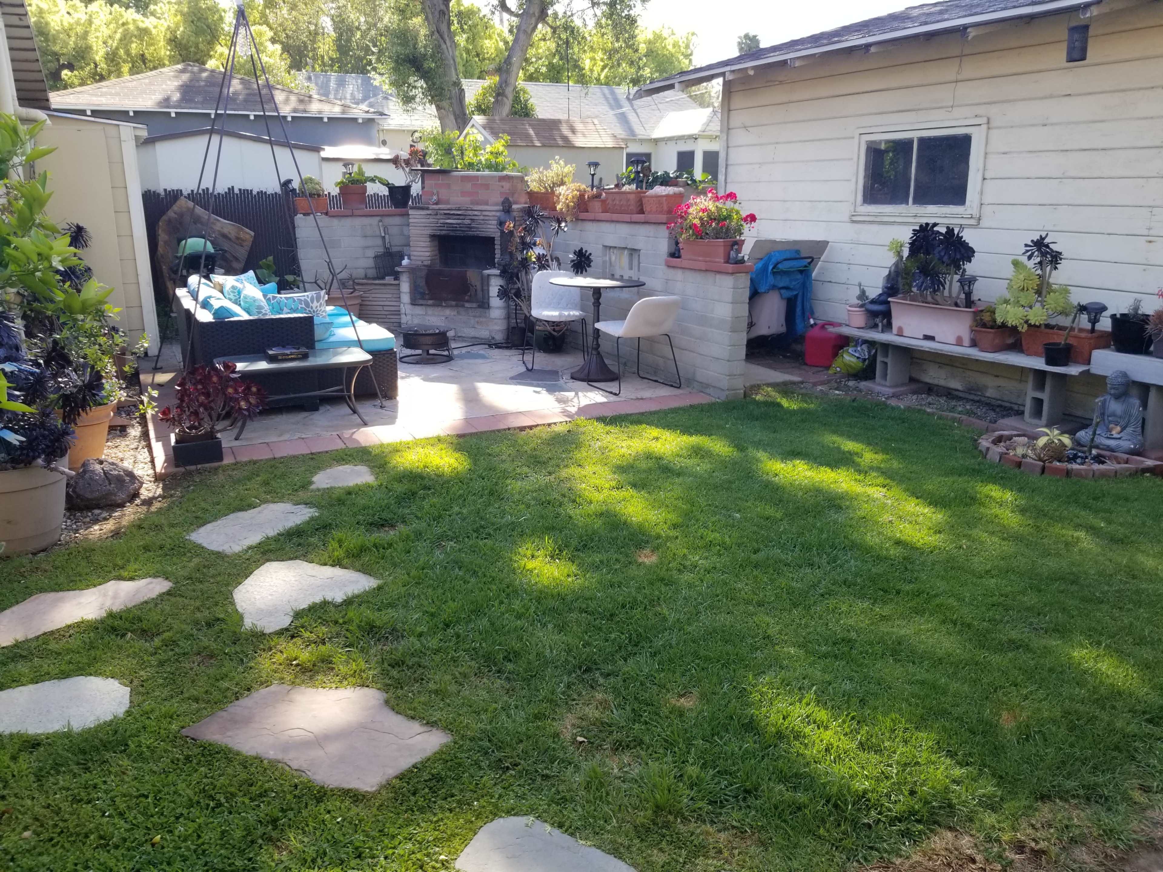 The image shows a backyard with a stone pathway leading to a seating area surrounded by potted plants and a grill.