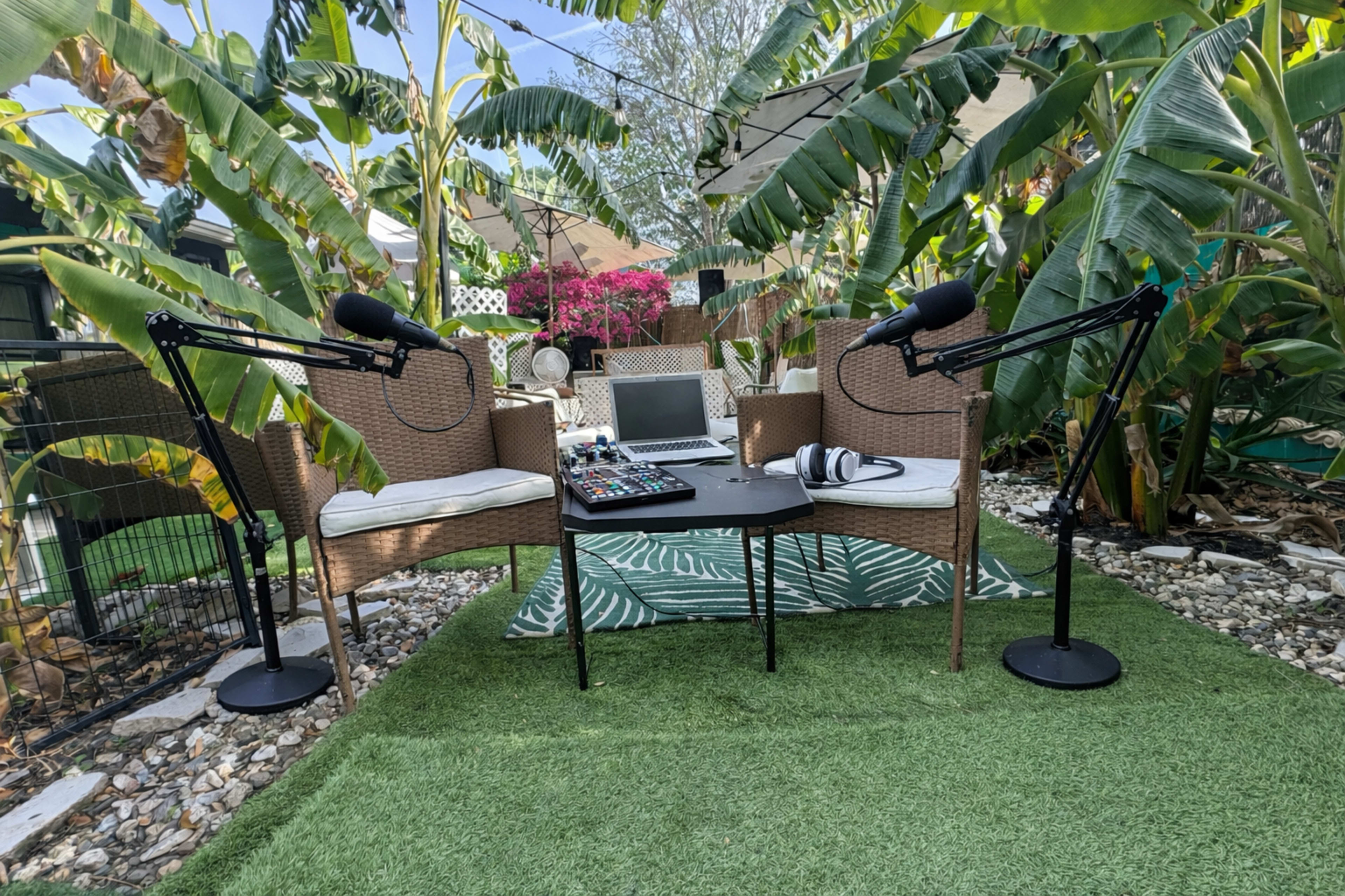 The image shows a podcast setup with two microphone stands and a control mixer placed on a table surrounded by chairs, tropical plants, and a decorative rug in an outdoor setting.