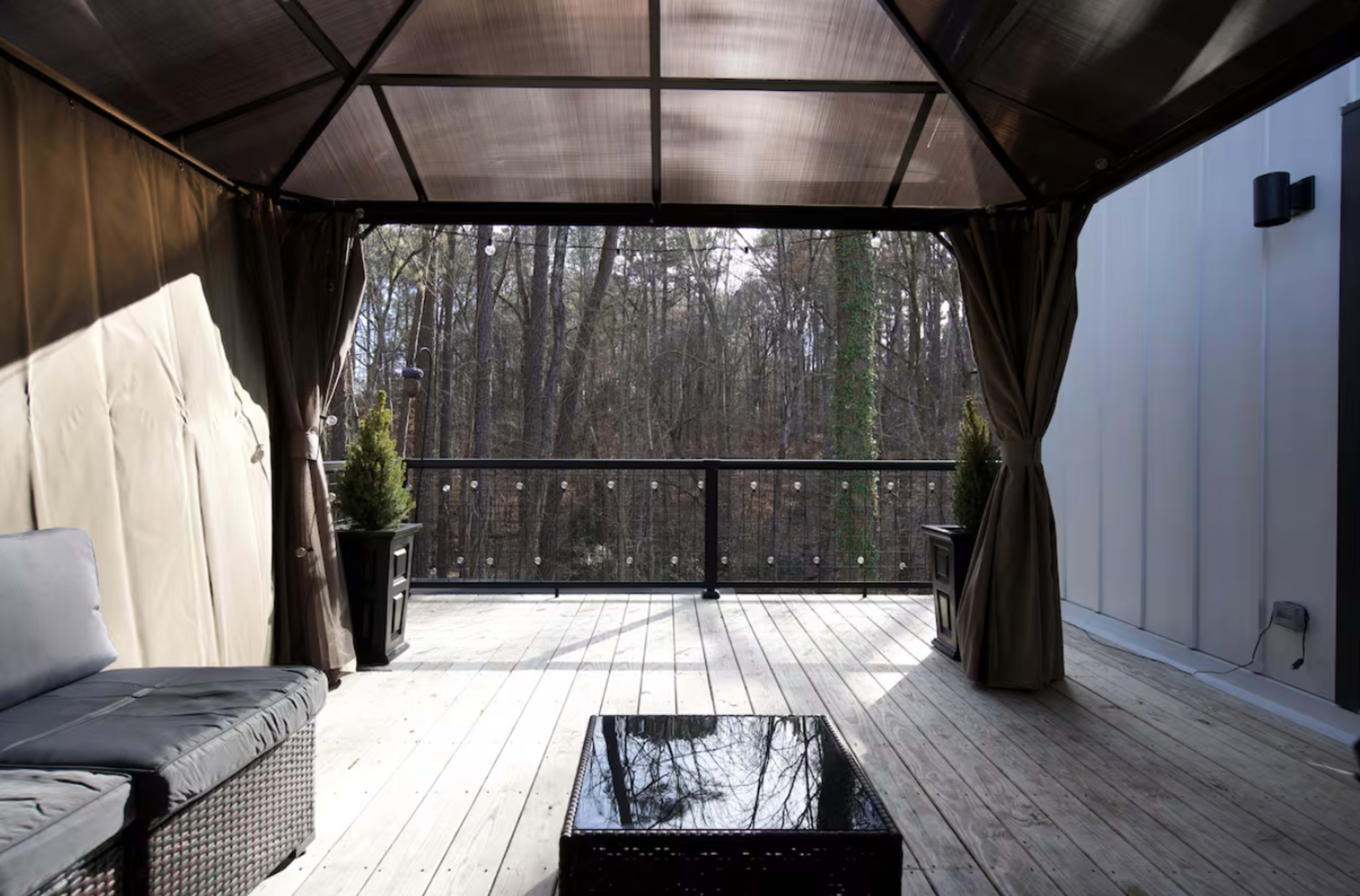 A shaded outdoor gazebo with seating, a glass table, and views of a wooded area beyond.