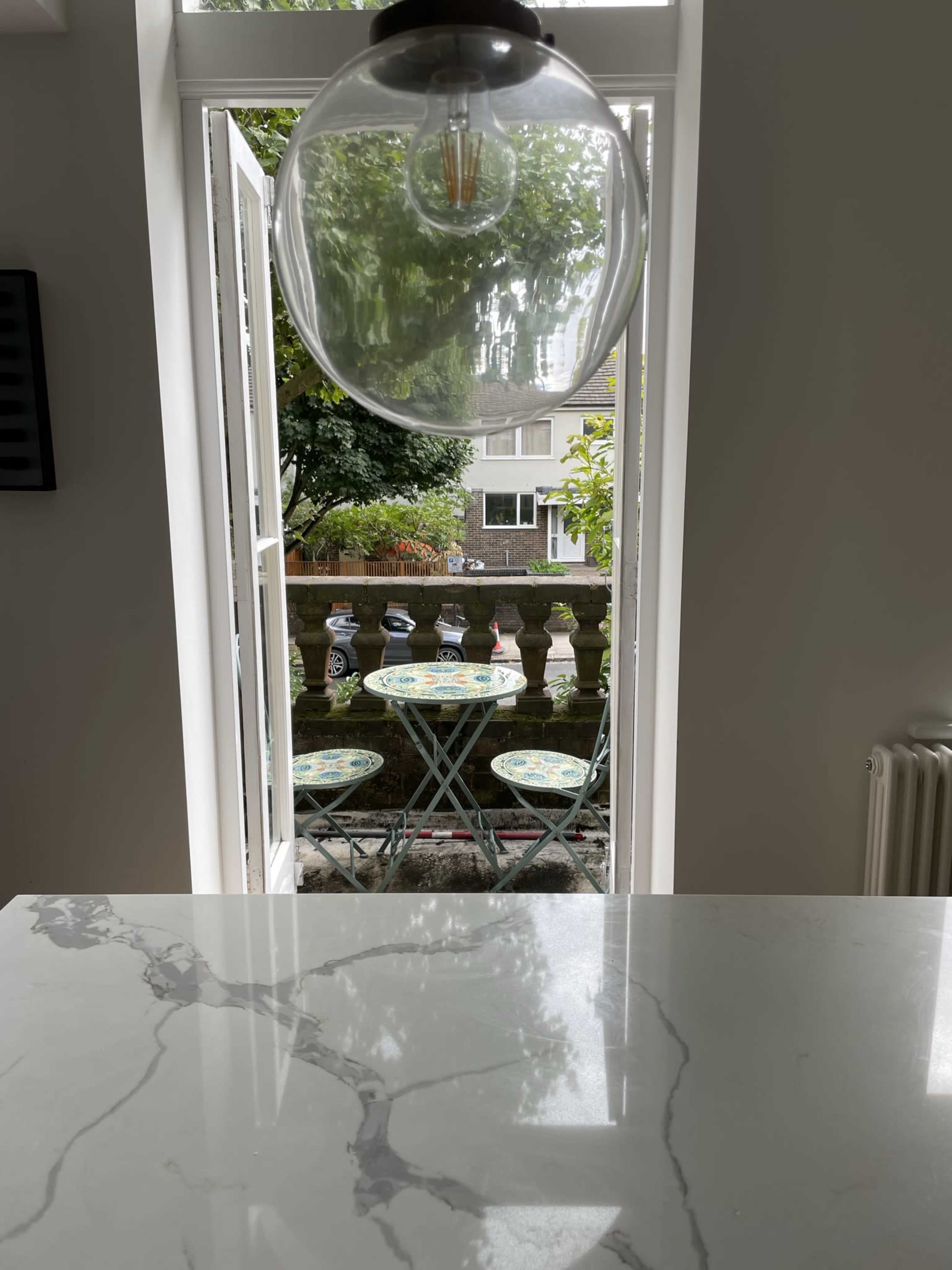 A round table with decorative chairs is set on a patio visible through an open window, framed by a large light fixture overhead.