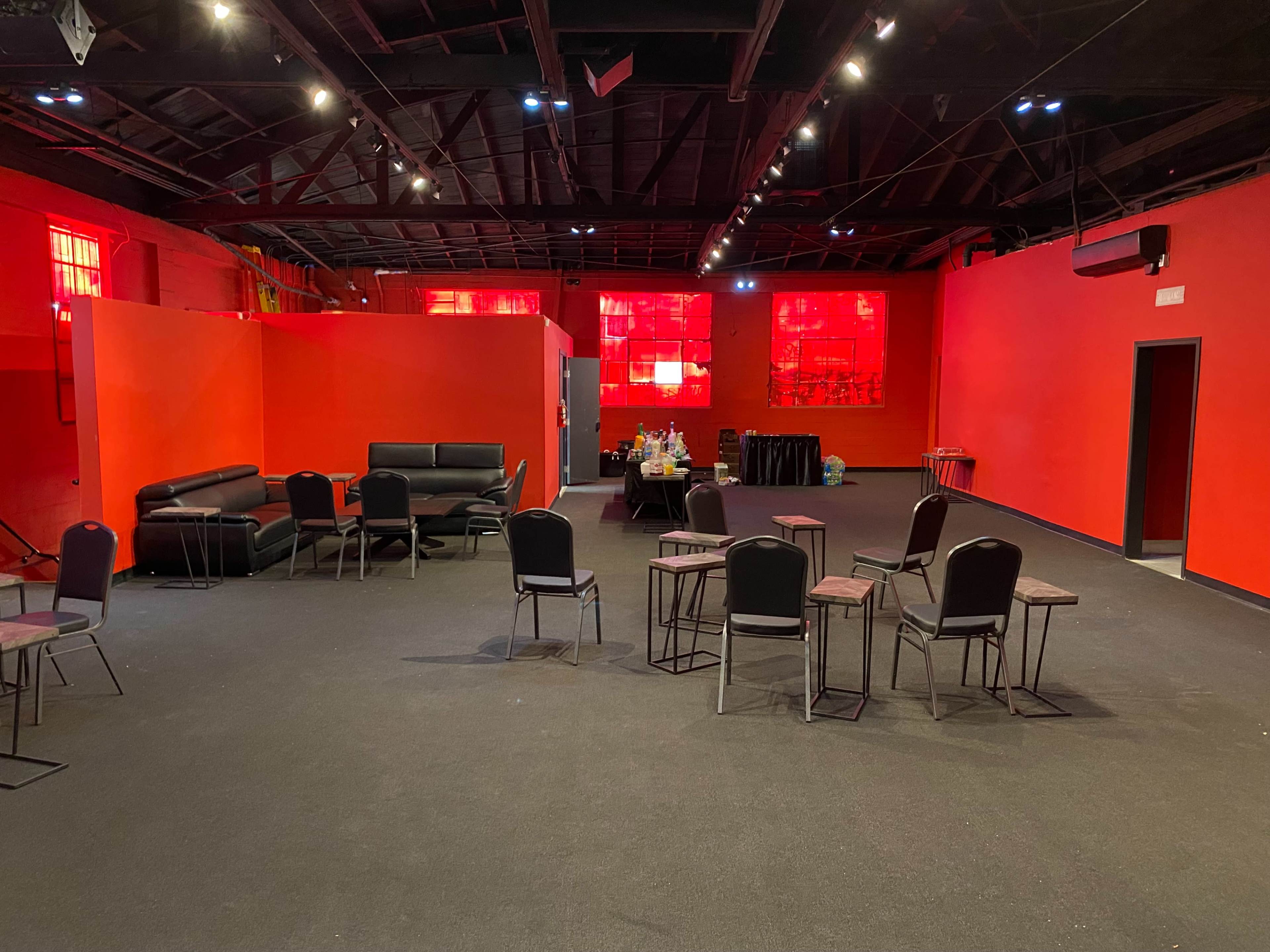 The image shows an empty room with bright red walls, black chairs arranged around the space, and a few scattered items, including a table and a couch.