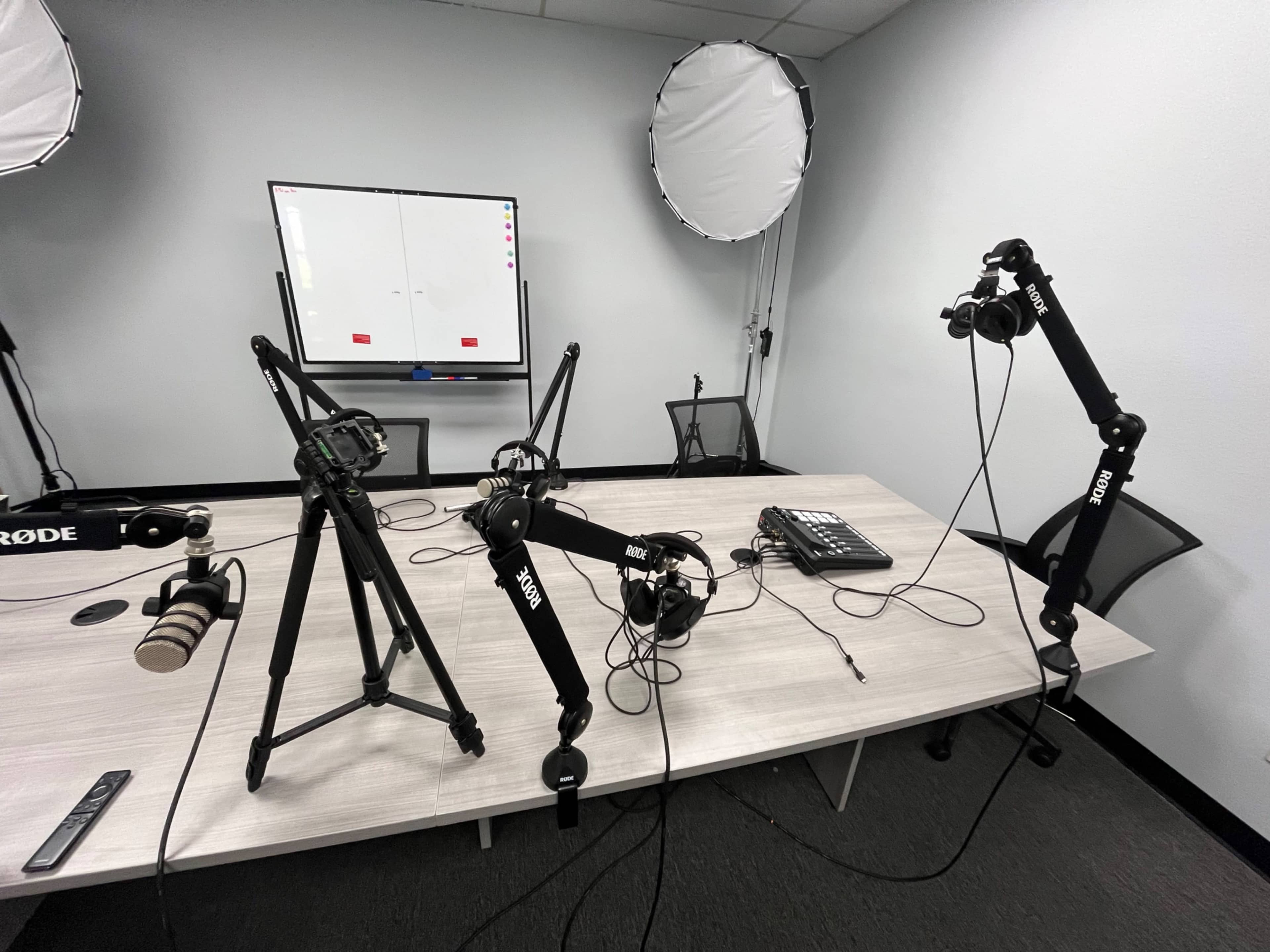 The image shows a conference room setup with a table, cameras mounted on tripods, and lighting equipment positioned around a blank whiteboard.