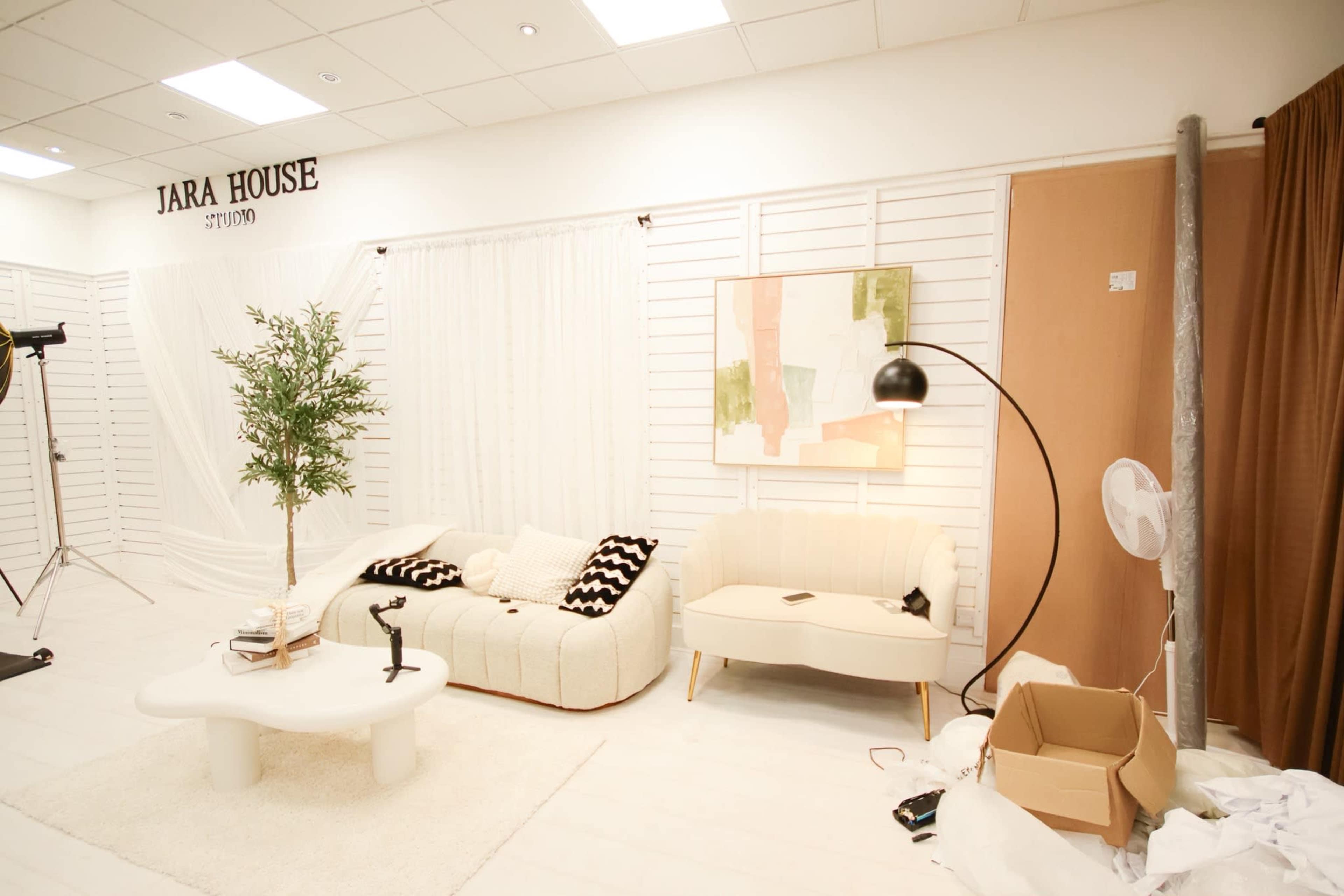 The image shows a bright studio space featuring a white sofa, a cream-colored couch, an abstract painting on the wall, and a potted plant, along with some studio equipment and a cardboard box in the corner.