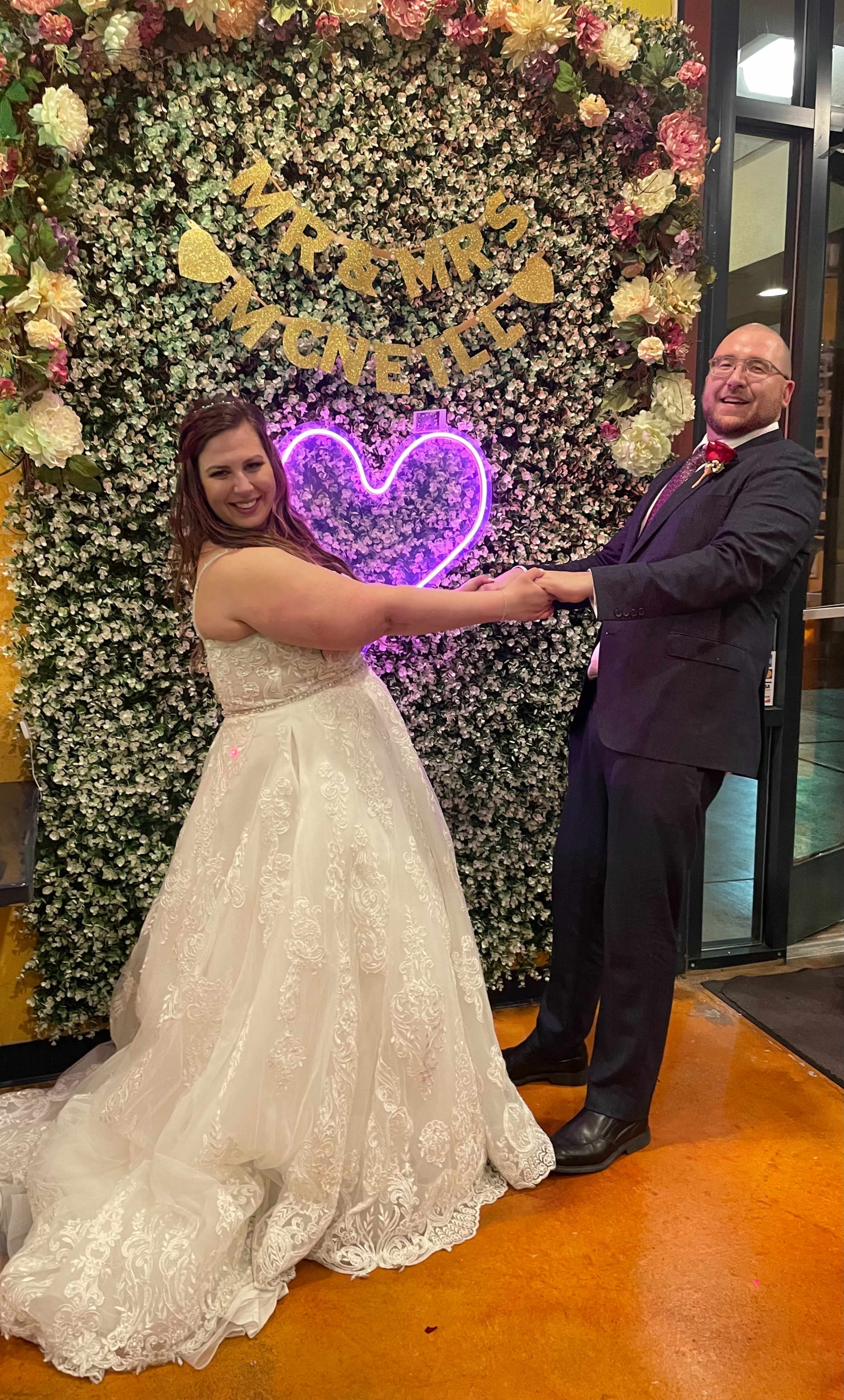 A bride and groom stand together in front of a floral backdrop featuring a neon heart and a sign that reads "MRS. MRS. AGNELLI."