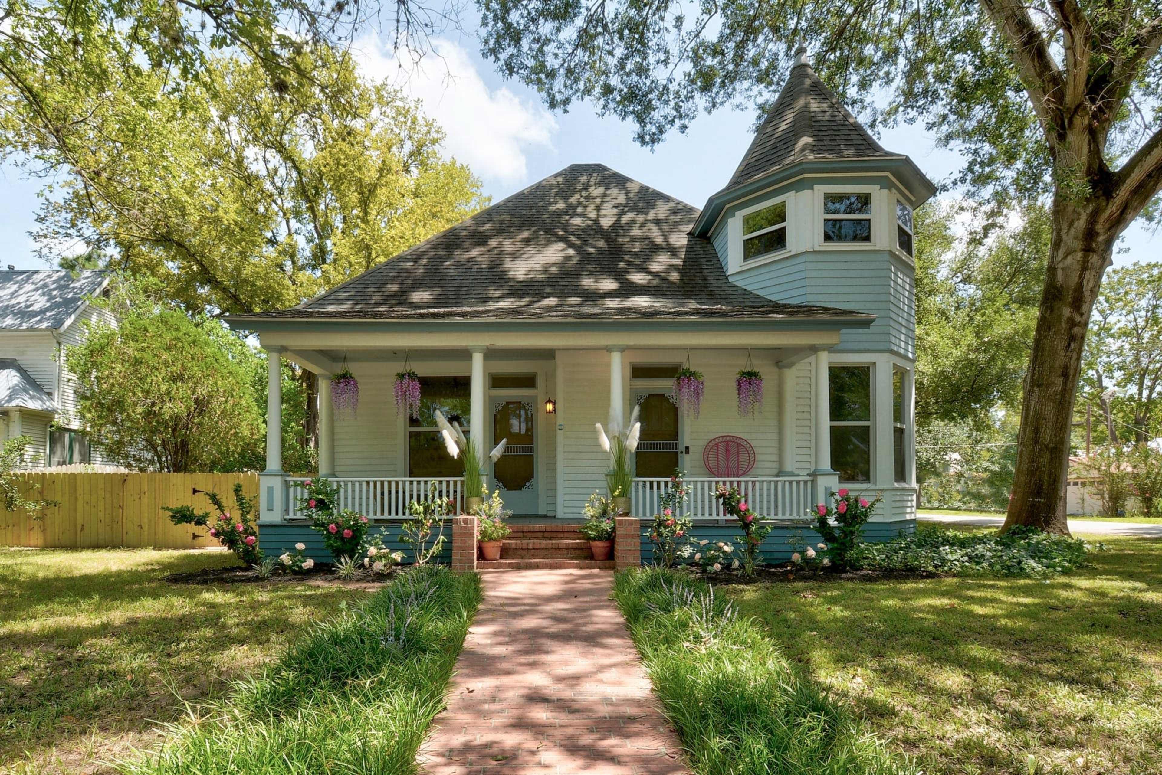 A light blue Victorian-style house with a turret and a front porch, surrounded by green grass and flower beds, features a brick pathway leading to the entrance.