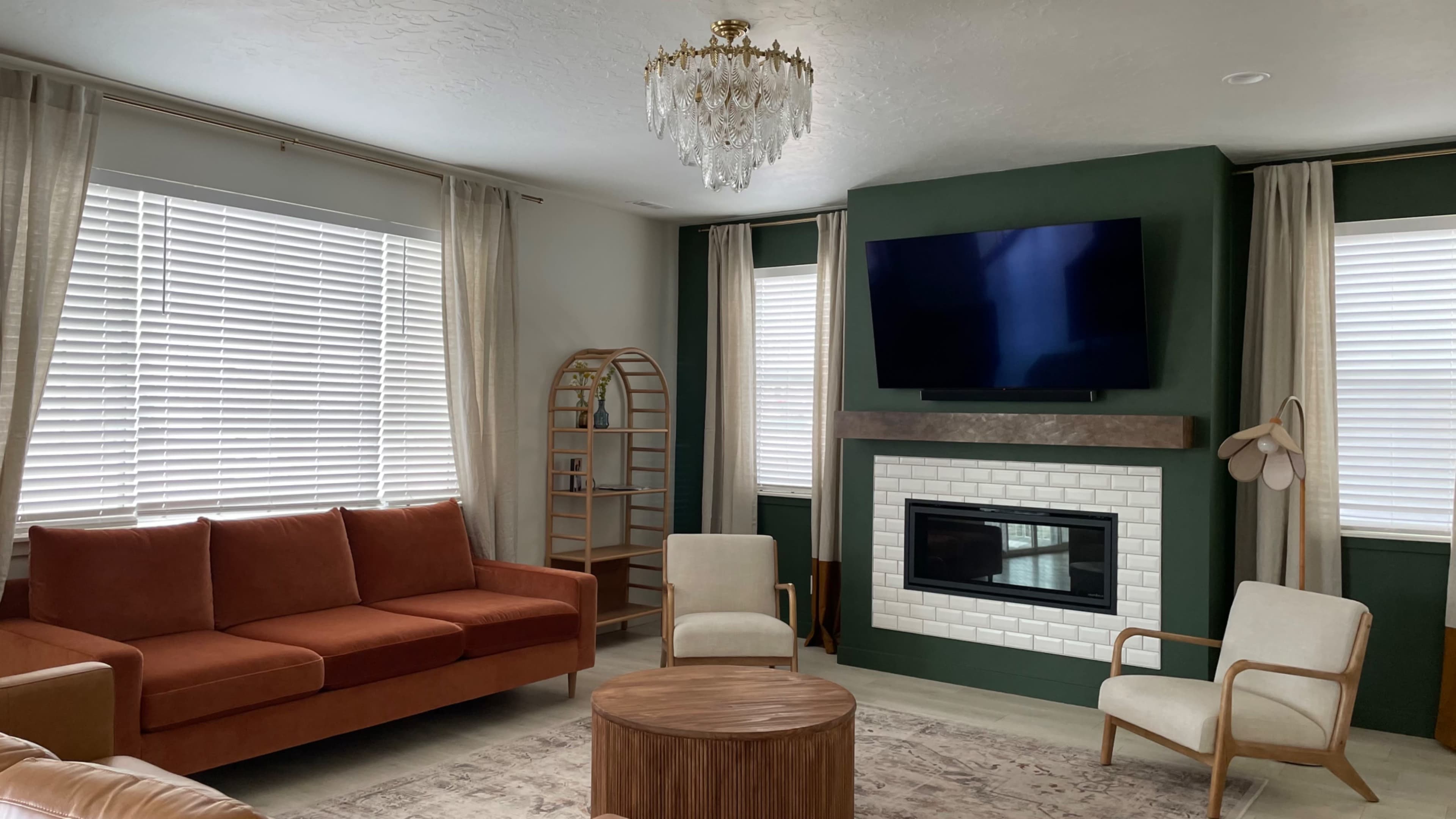 A living room with a green accent wall, a mounted television, a white fireplace, and a mix of upholstered furniture including an orange sofa and cream-colored chairs.