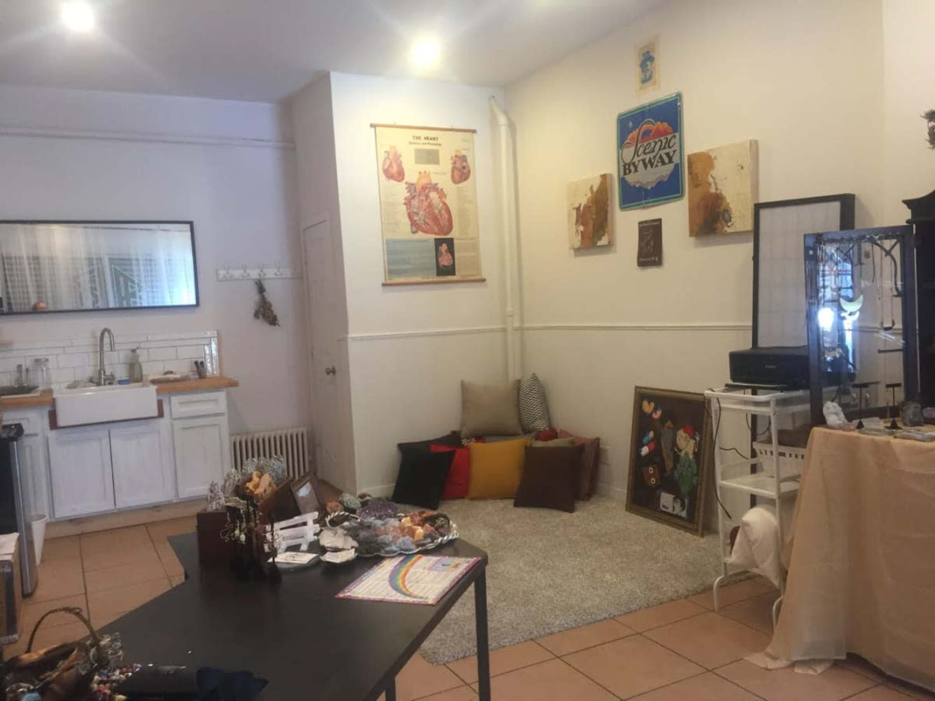 A small, softly lit room with a seating area on a rug, a black display stand, and a wooden table cluttered with various items.