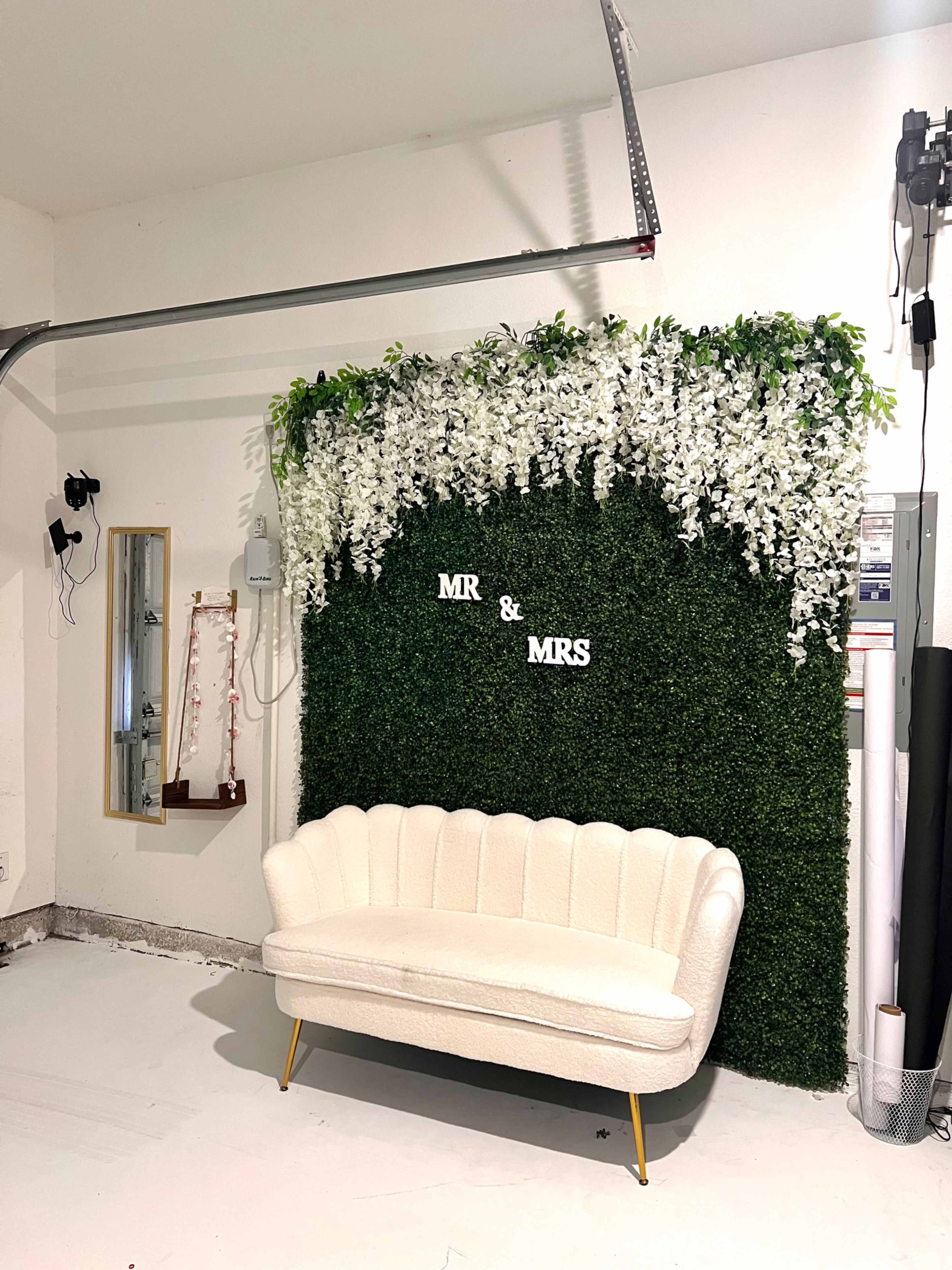 The image shows a white couch positioned in front of a decorative green wall adorned with white flowers, displaying the words "MR & MRS."