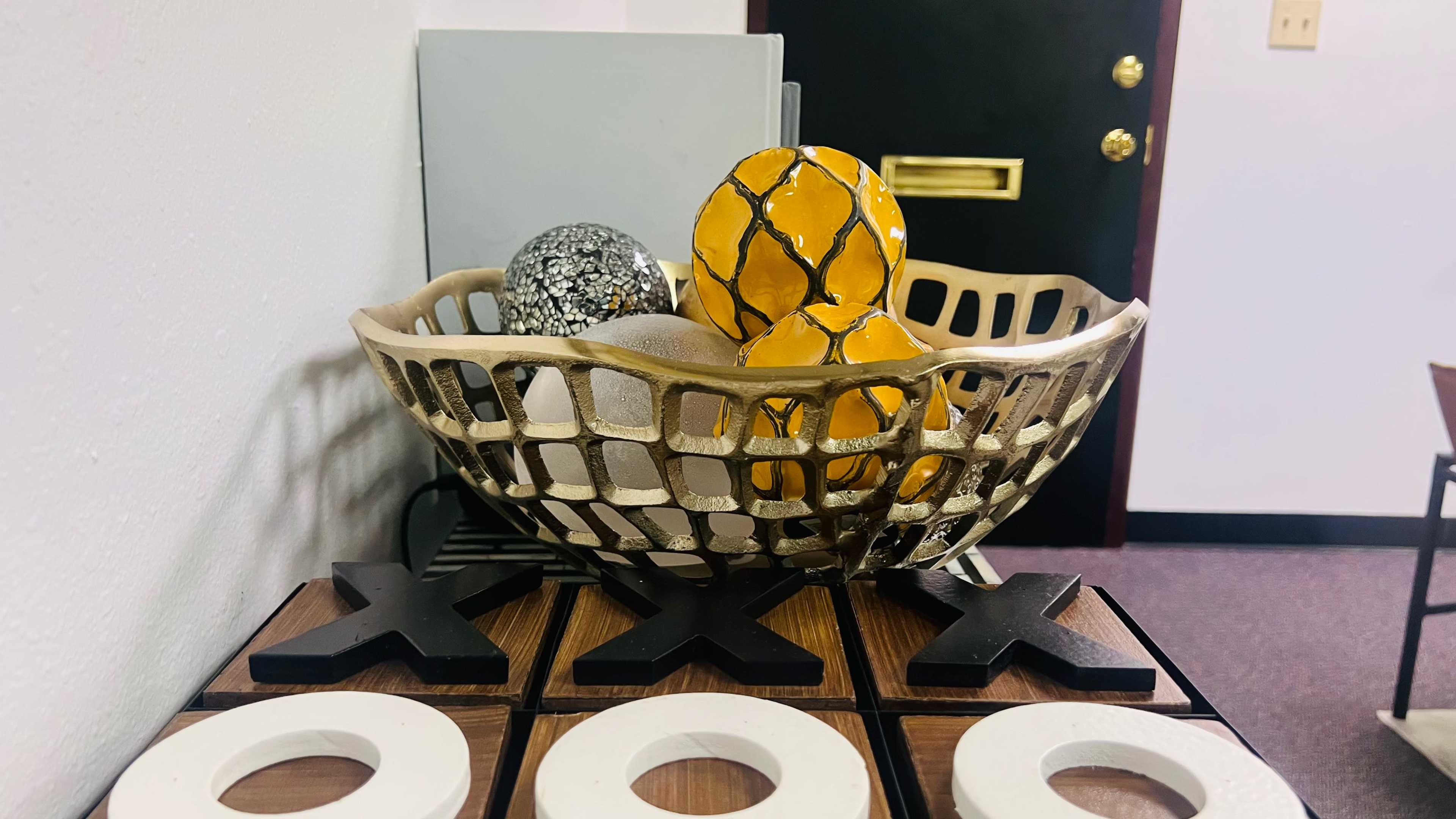 A decorative bowl filled with ornamental balls rests on a wooden surface featuring a tic-tac-toe game with black and white pieces.