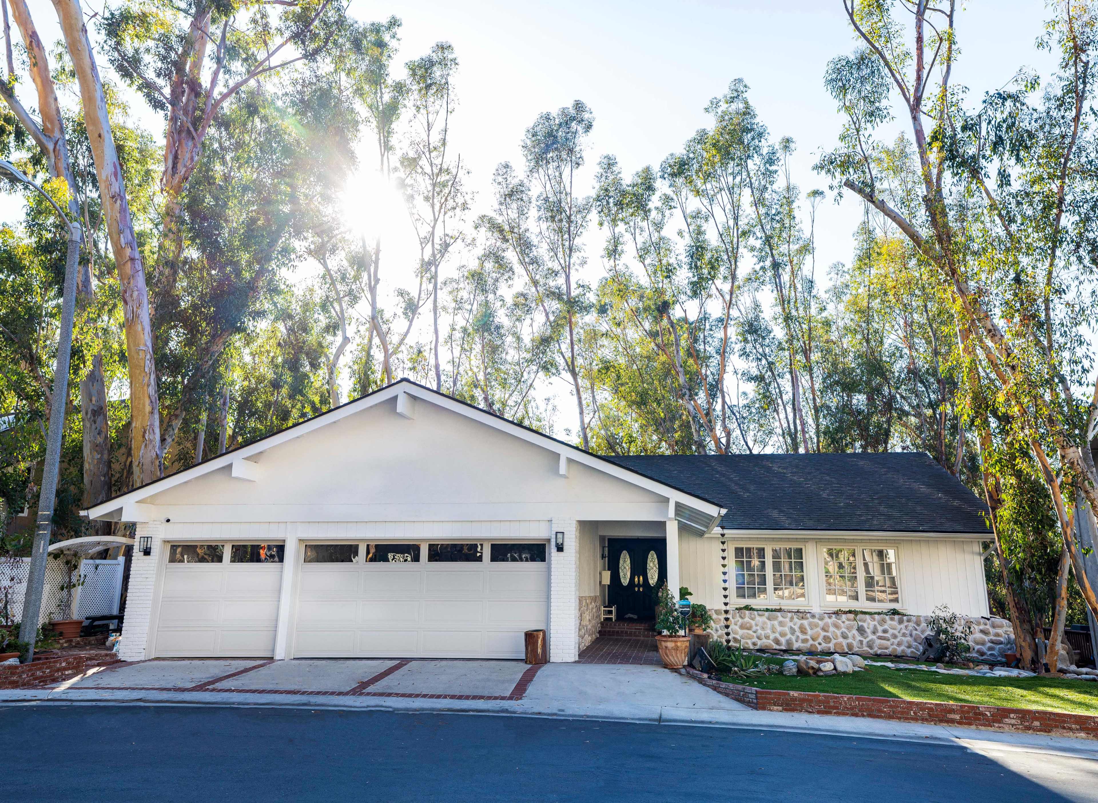 A single-story white house with a stone accent wall and three garage doors is surrounded by tall trees.