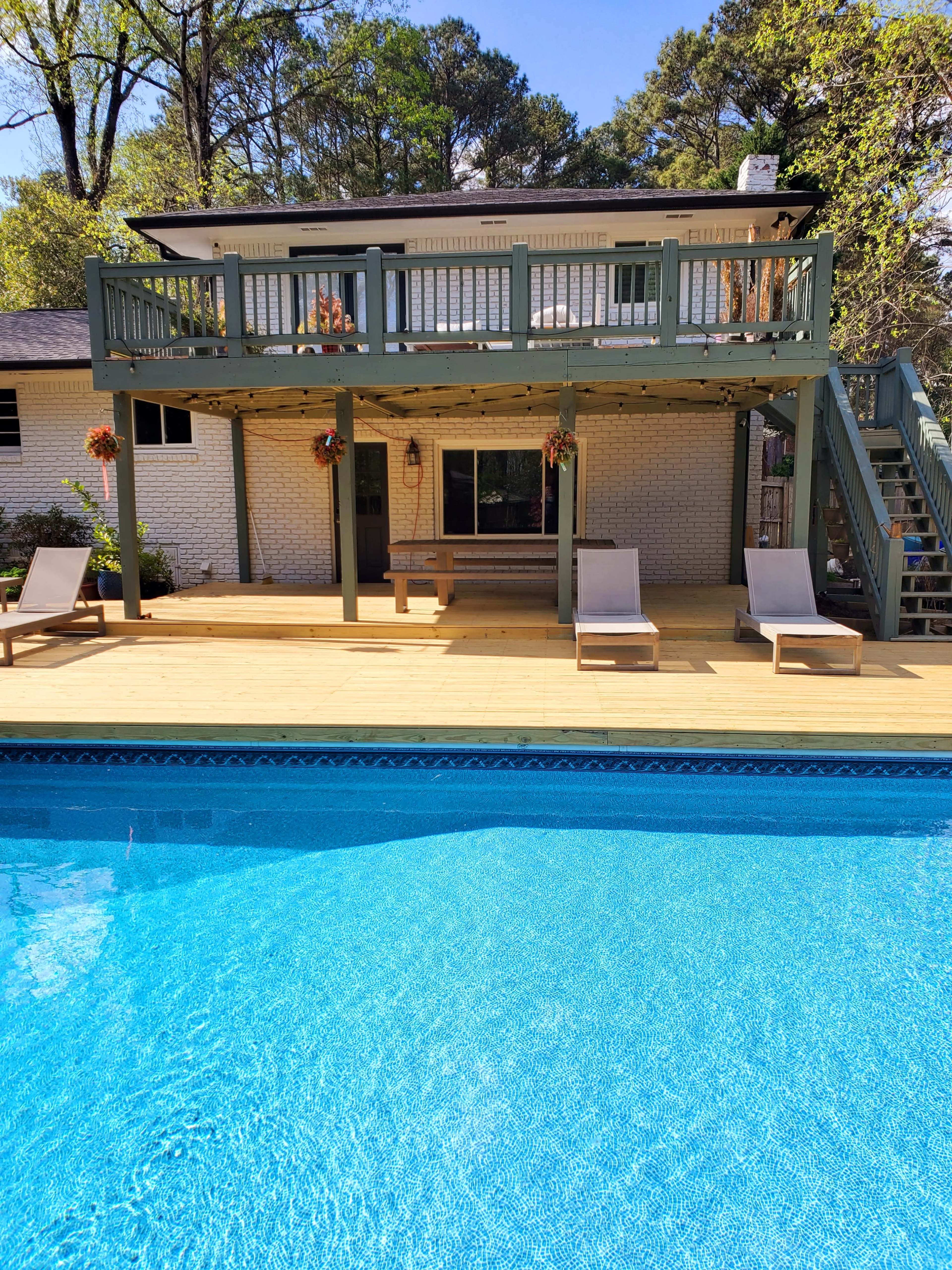 The image shows a two-story house with a wooden deck overlooking a swimming pool, featuring lounge chairs beside the pool and a staircase leading to the upper level.