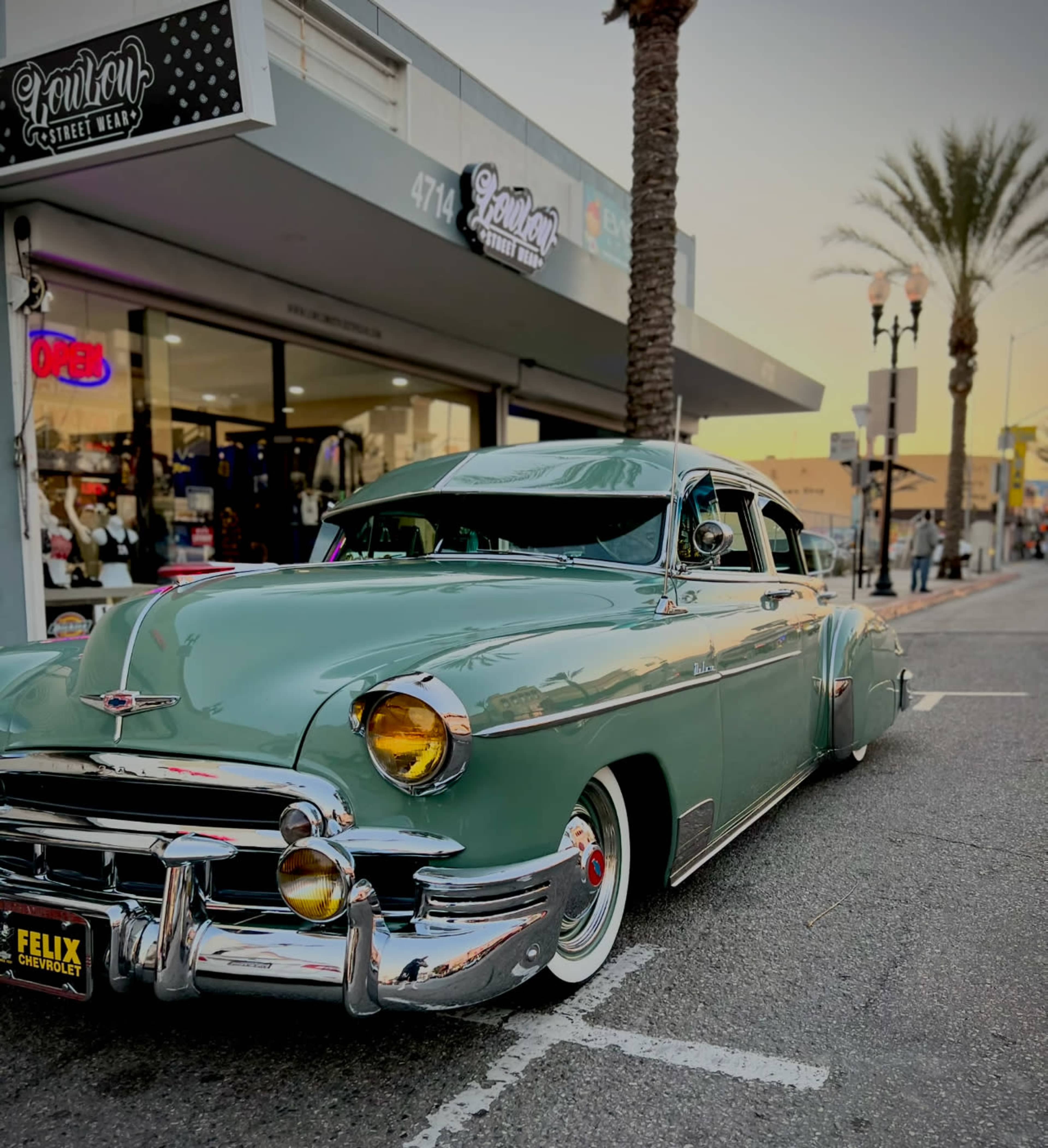 A classic green Chevrolet car is parked on a city street near a storefront and palm trees.