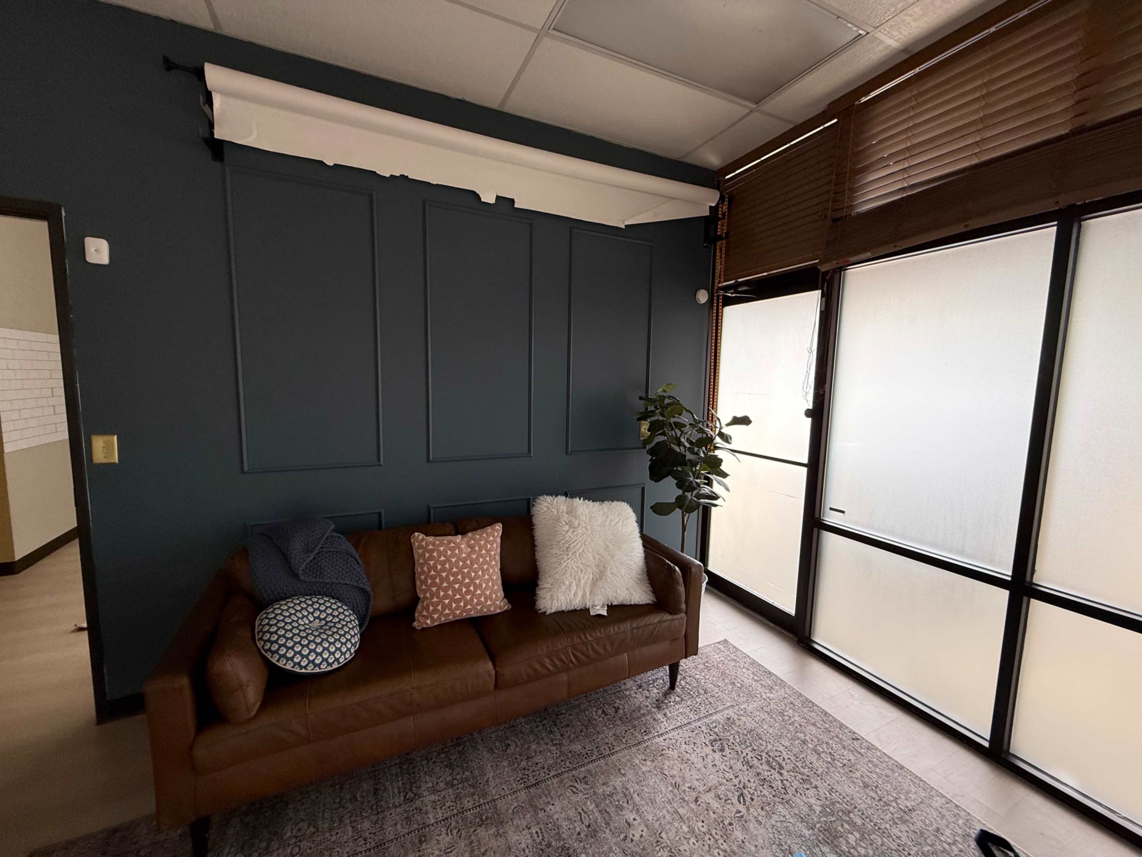The image shows a modern living room with a brown leather sofa, decorative pillows, a potted plant, and large windows filtered by blinds.