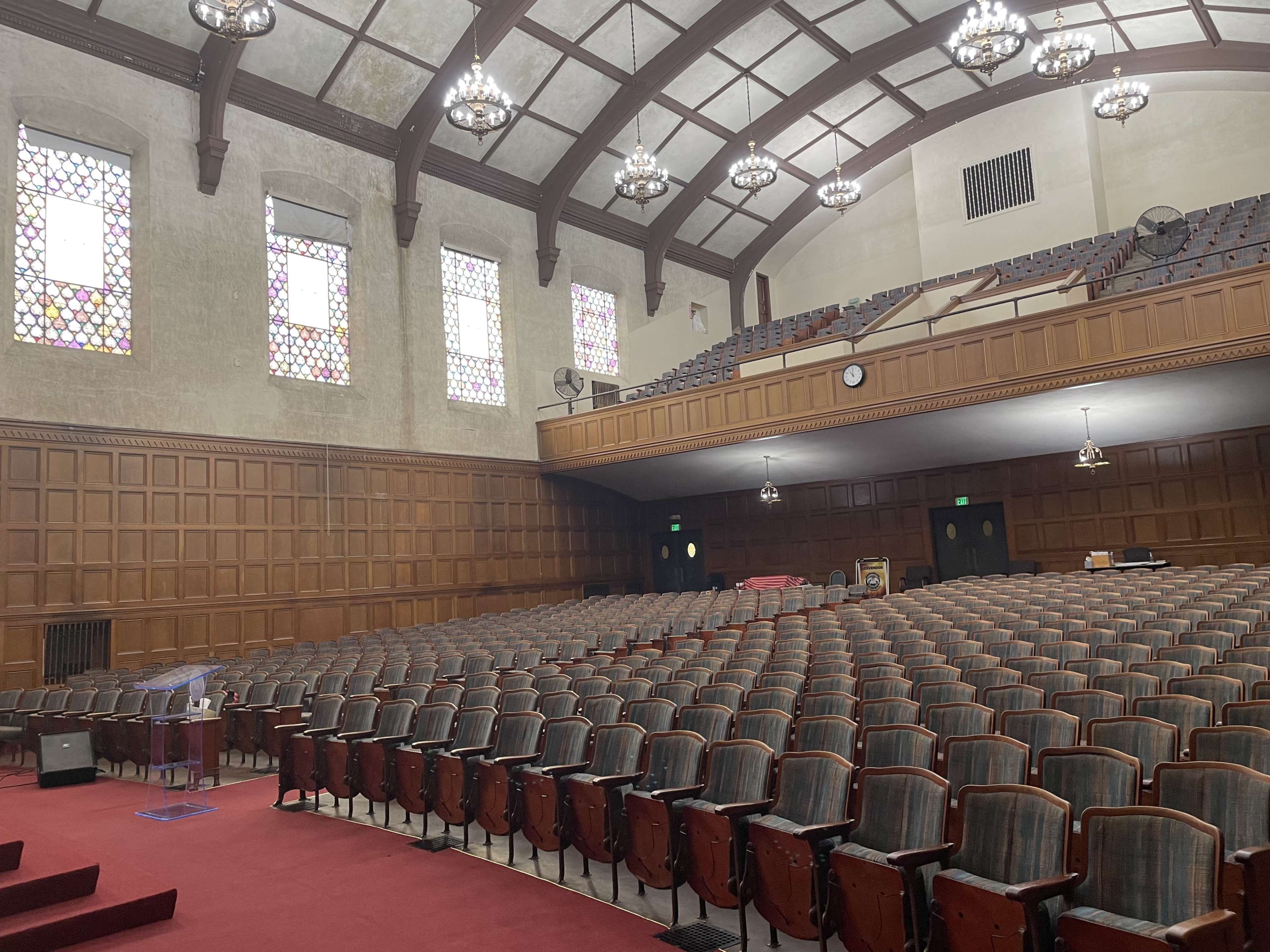 The image shows a large, empty auditorium with wooden seating, stained glass windows, and a high, vaulted ceiling.