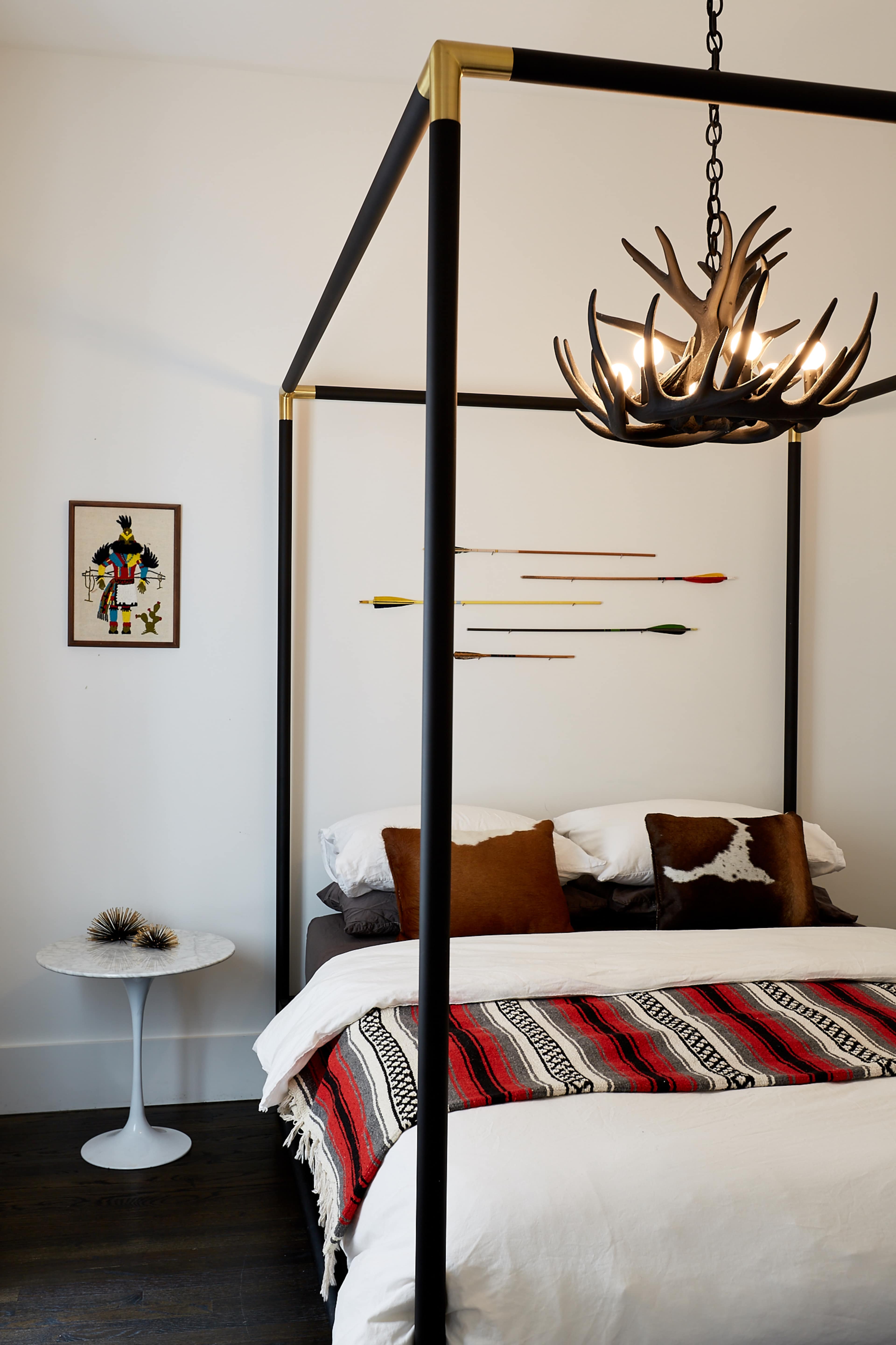 A modern bedroom features a four-poster bed with a chandelier made of antlers, decorative arrows on the wall, and a side table next to the bed.