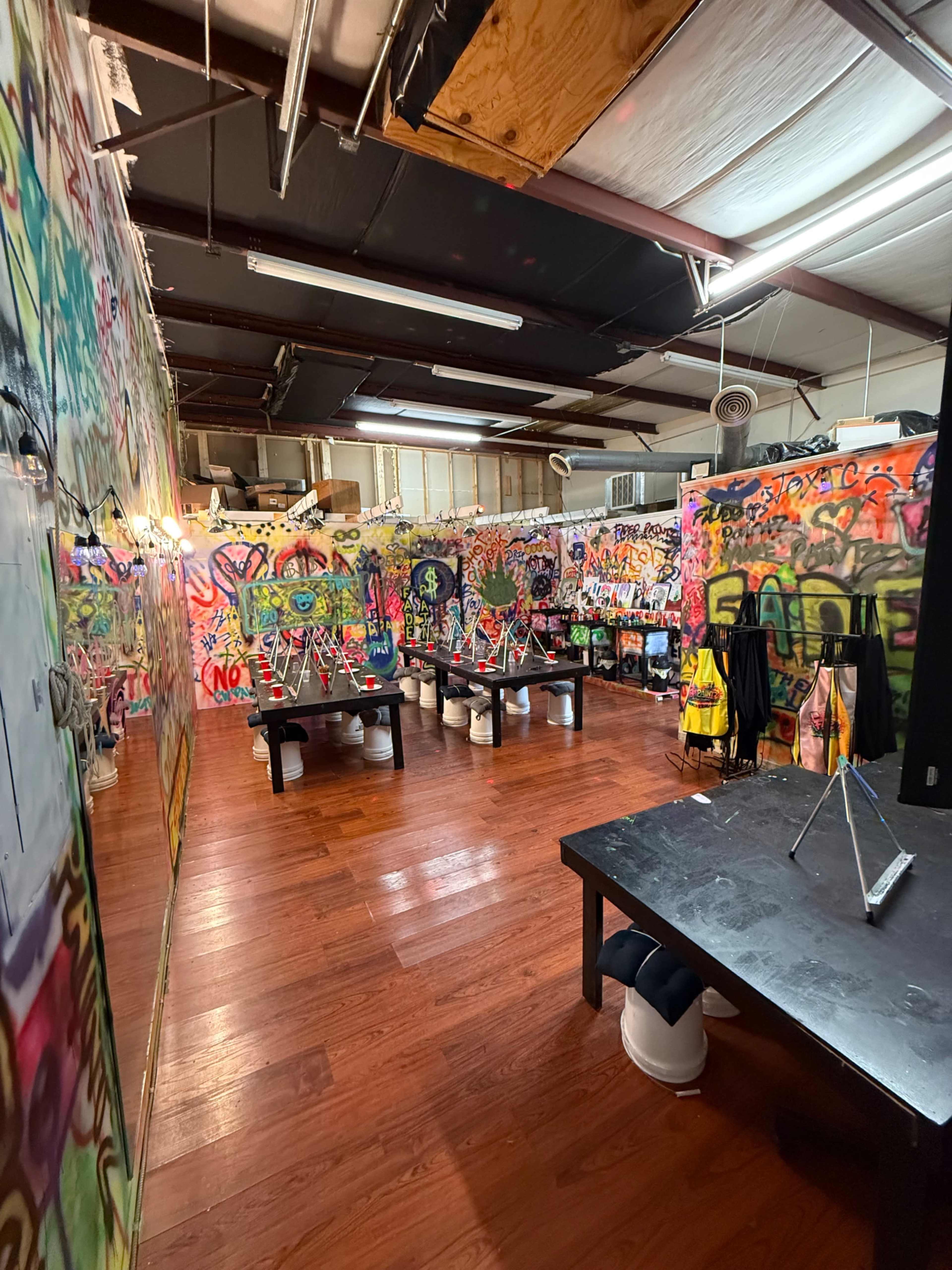 The image shows an indoor space with walls covered in colorful graffiti, featuring several black tables and various art supplies scattered around.