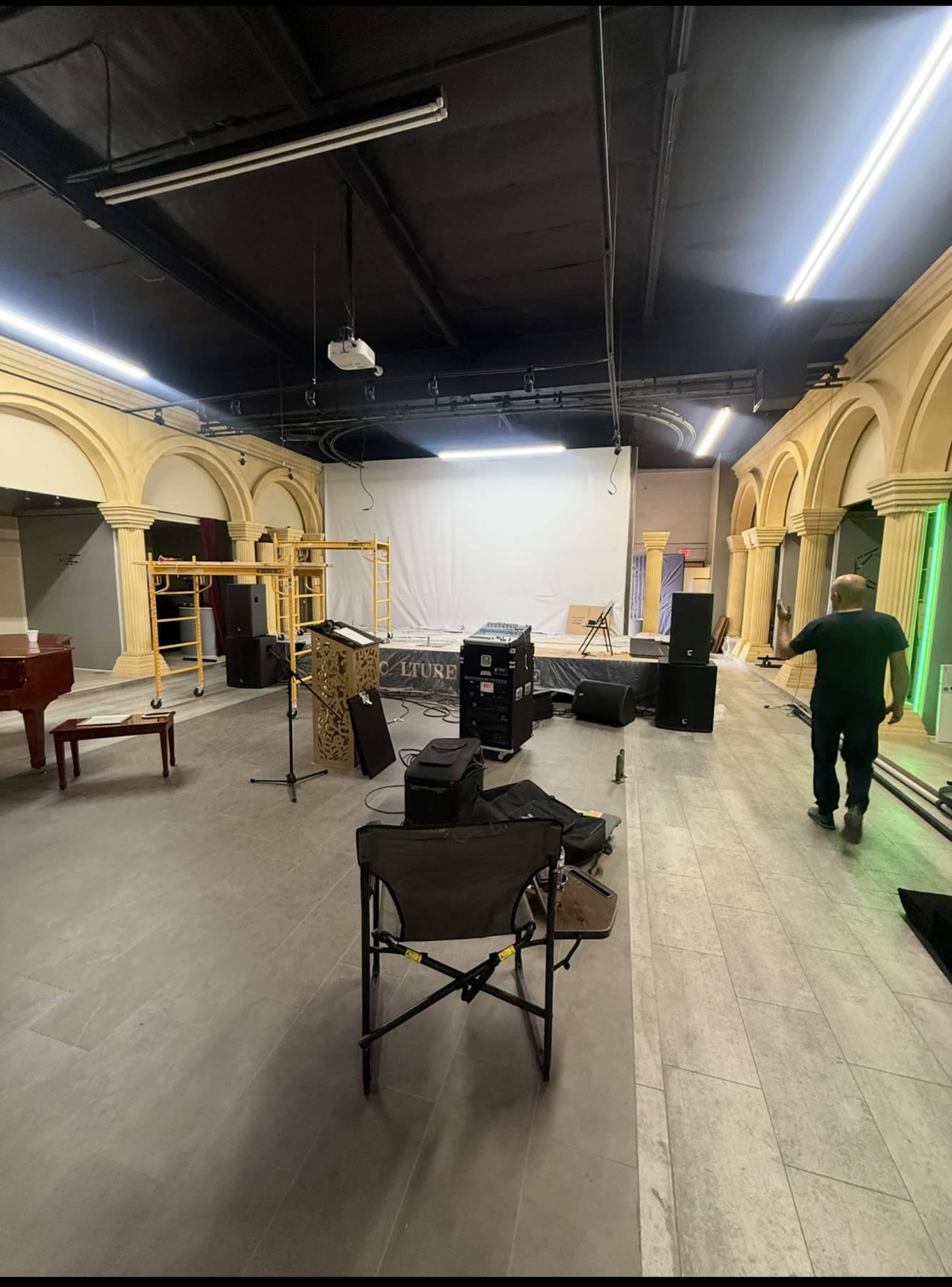 The image shows a spacious venue with a stage setup, including a grand piano, speakers, and lighting equipment, while a man walks towards the back.