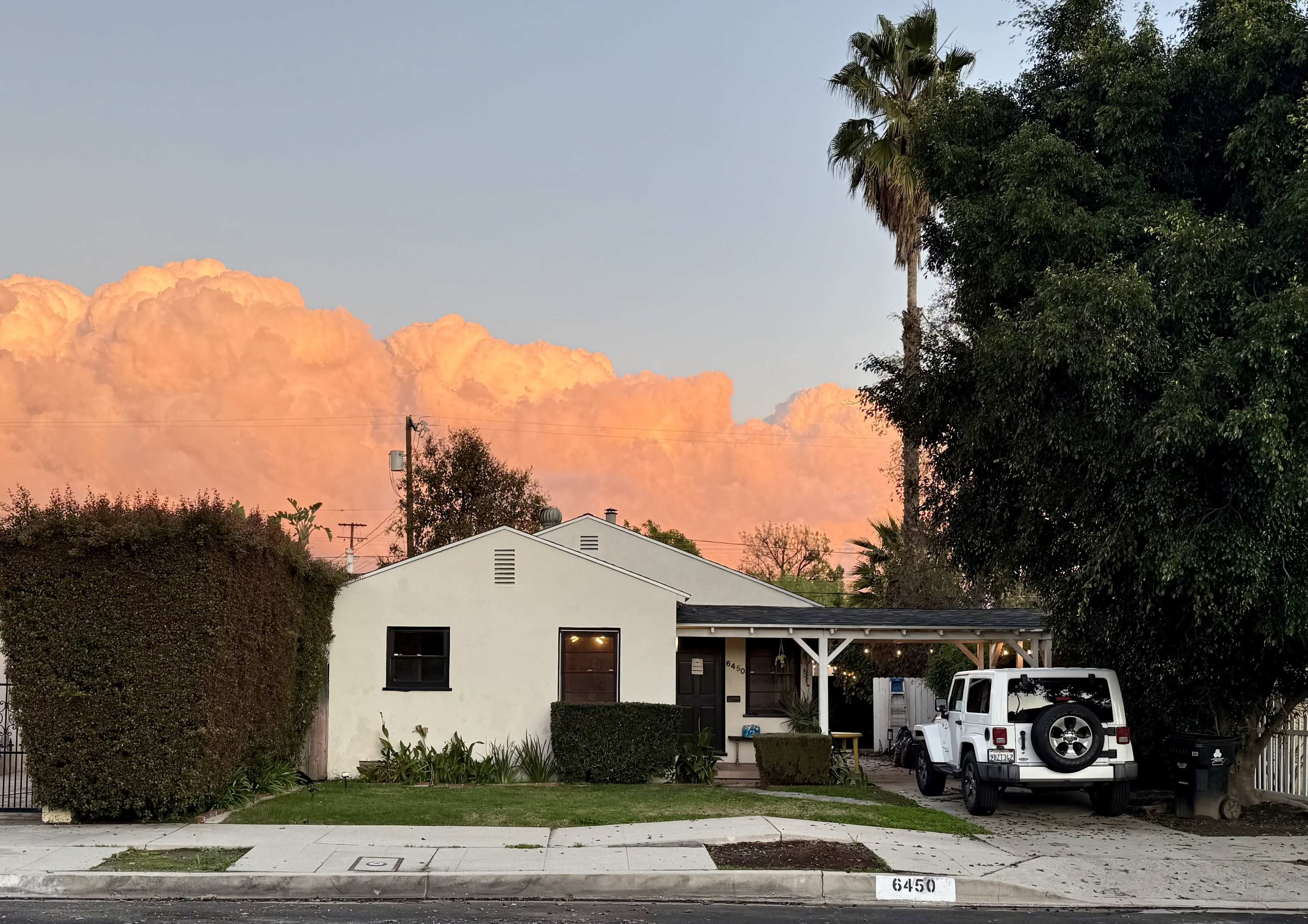 A single-story house with a white exterior, a front porch, and a Jeep parked on the driveway is situated under a colorful sunset sky.