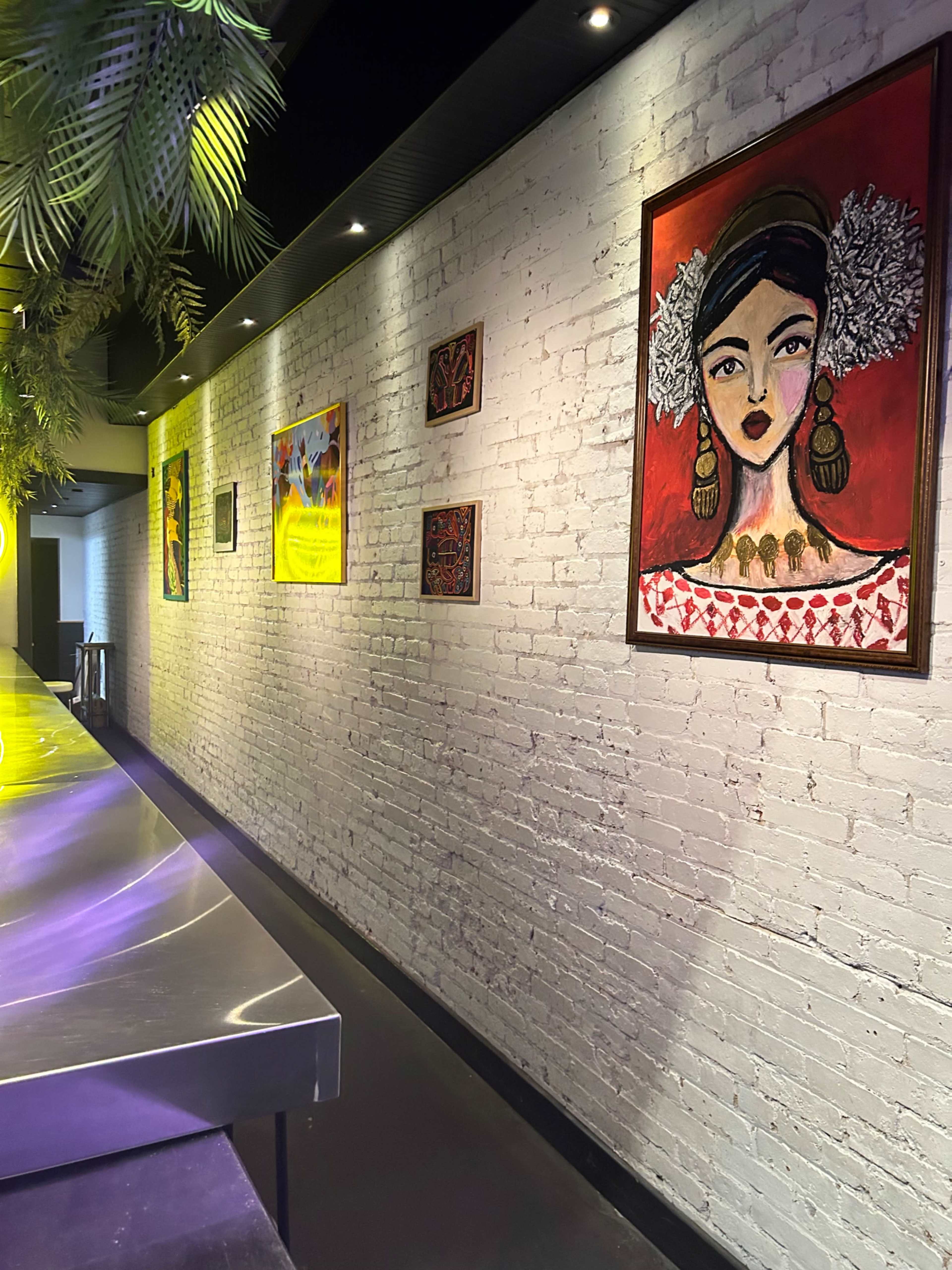 A long, brightly lit wall displays various colorful paintings alongside a metallic counter in a modern space.