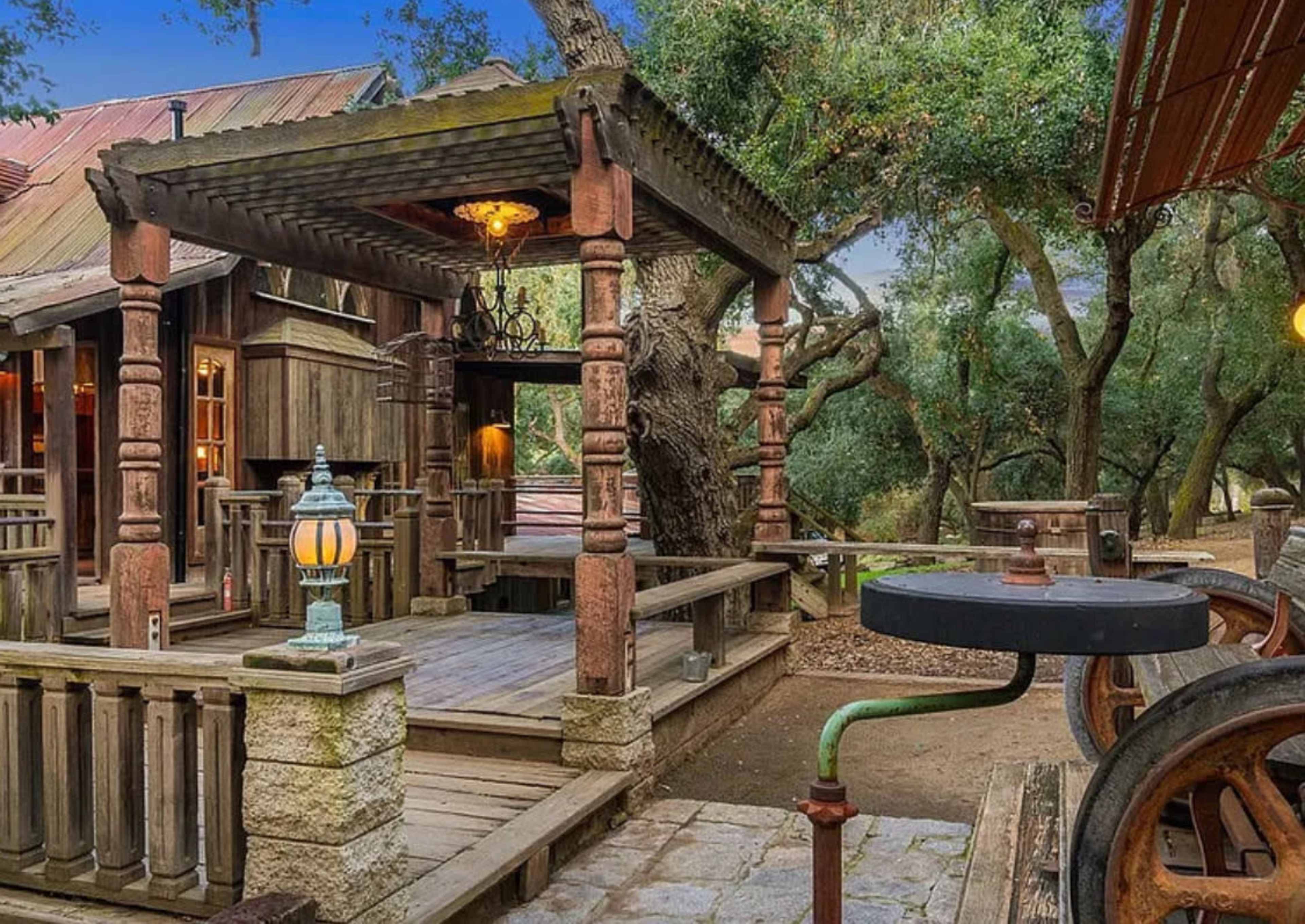 The image shows a rustic wooden deck area with a pergola supported by carved columns, surrounded by trees and outdoor fixtures.