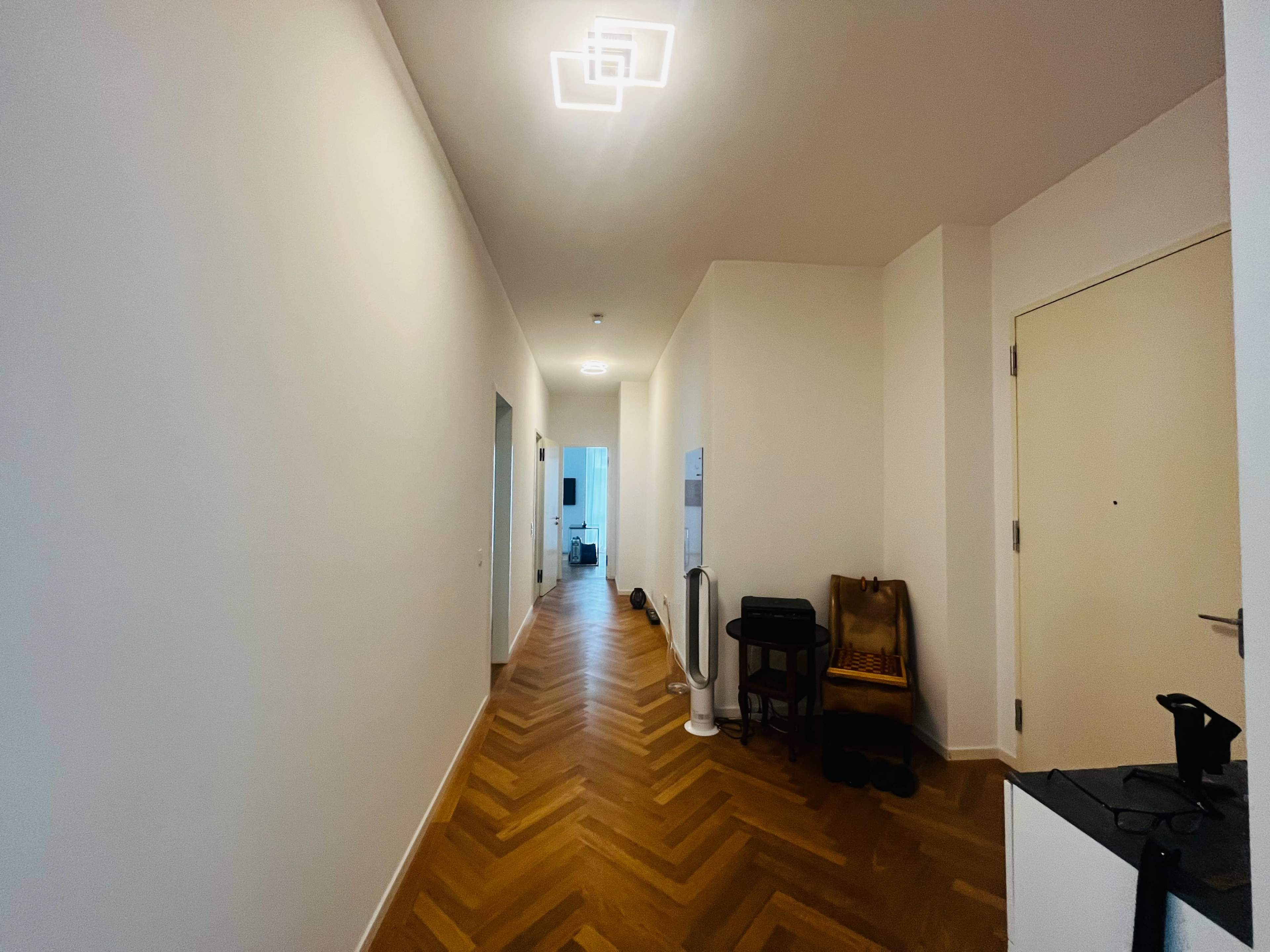 A long hallway with light-colored walls, a herringbone wood floor, and a ceiling light fixture at the far end.