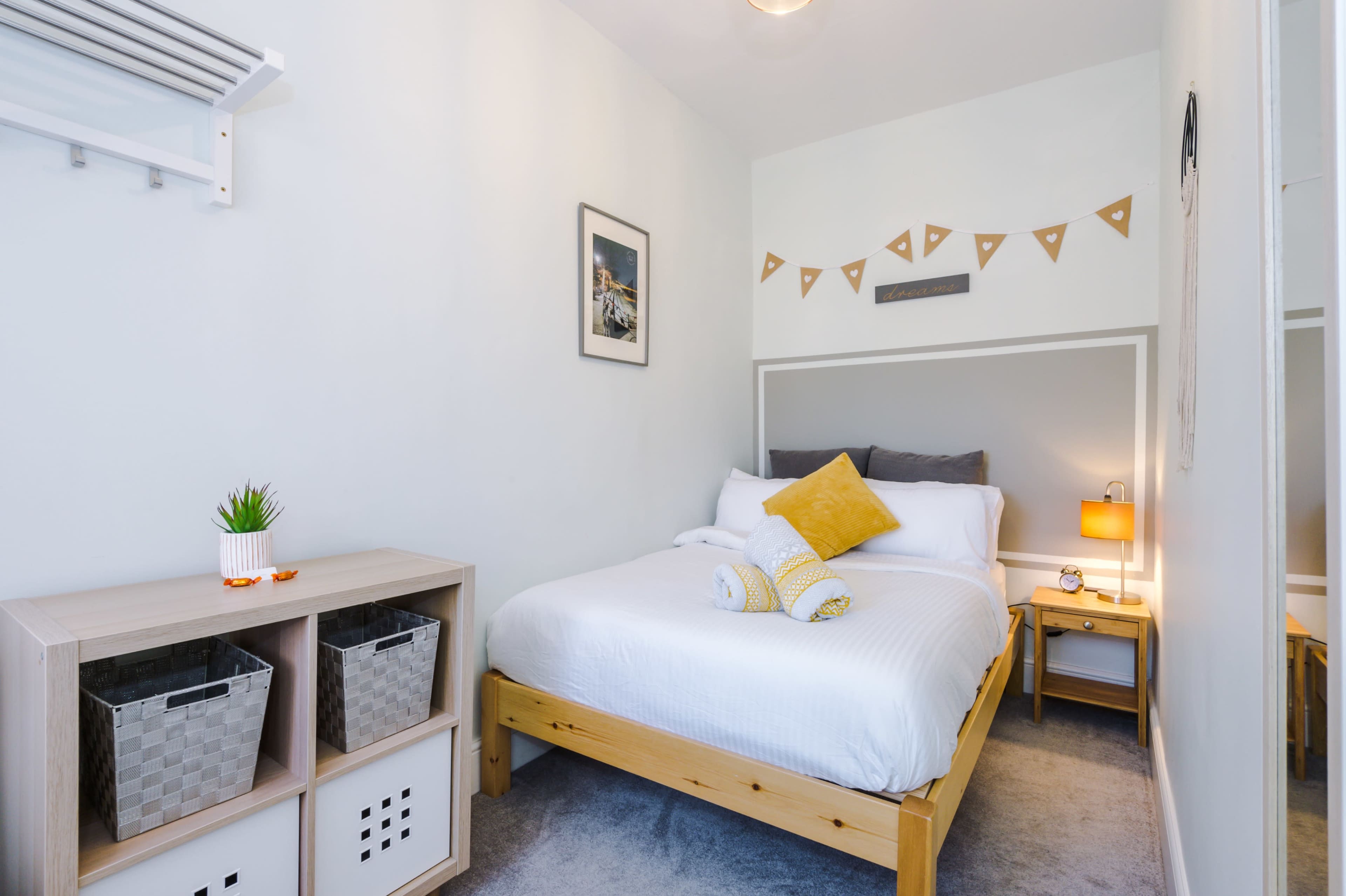 A small bedroom features a double bed with white linens, a nightstand with a lamp, and a decorative storage unit with baskets.